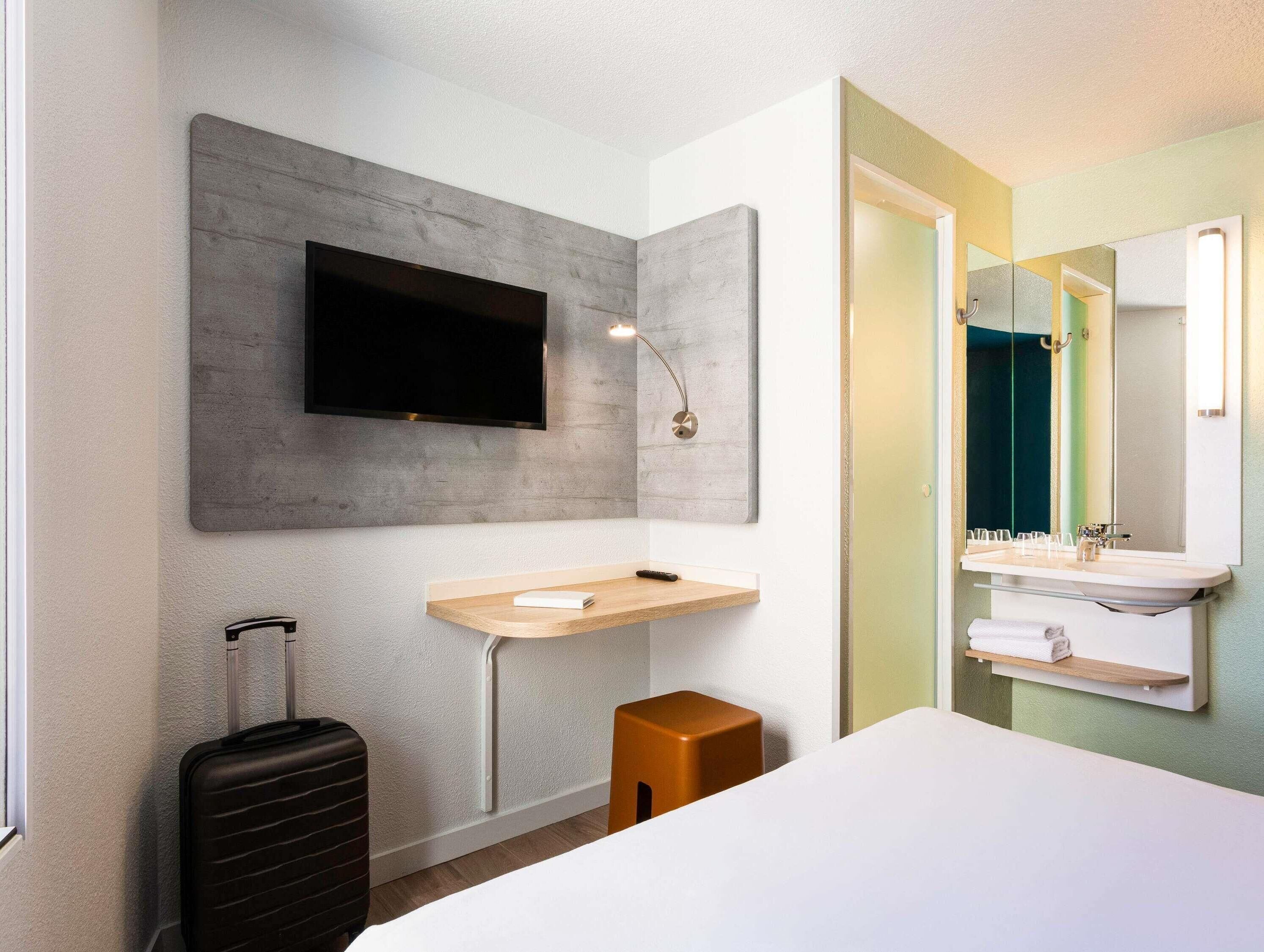 Ein modernes Hotelzimmer mit Flachbildfernseher an einer grauen Wand, einem Nachttisch aus Holz mit Lampe, einem kleinen Hocker, einem großen Bett und einem Badezimmer mit Spiegel, Waschbecken und Handtuchhalter.