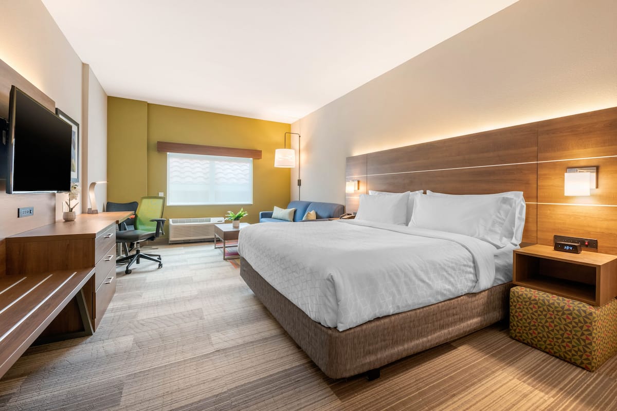 A hotel room with a large bed, a desk, a chair, a TV, and a window with blinds.