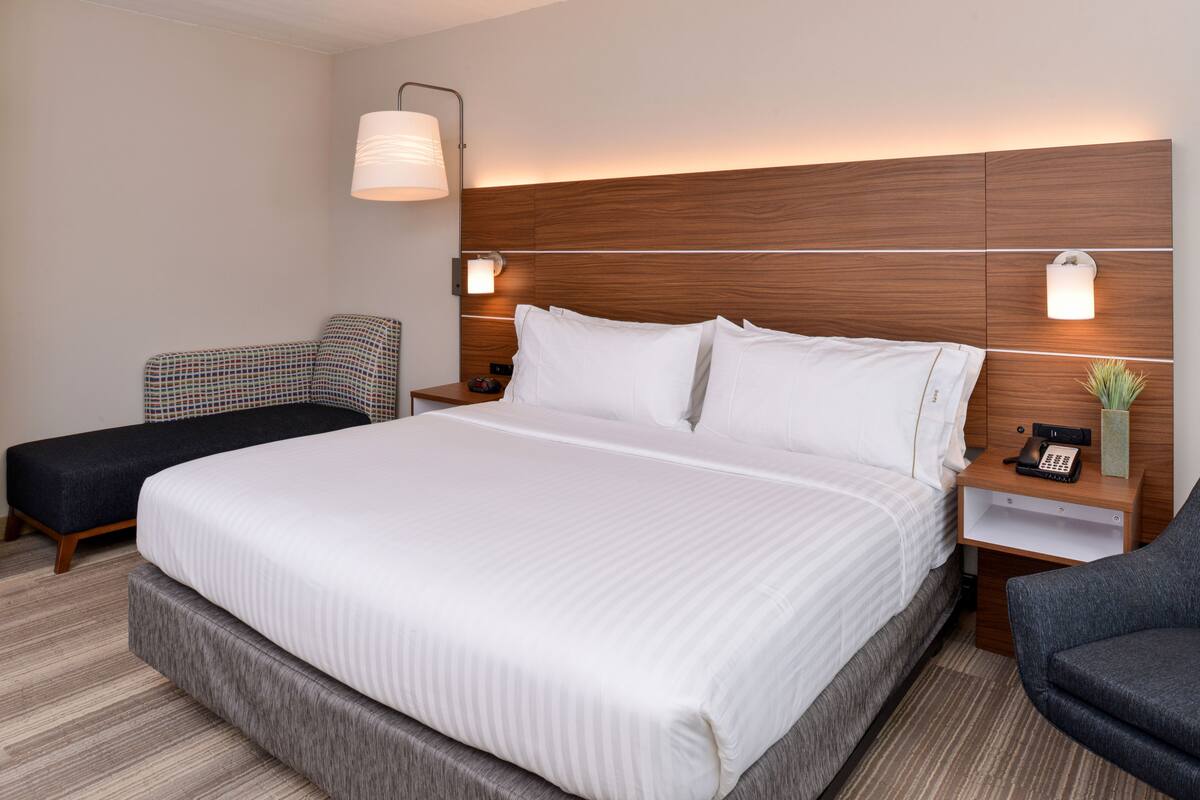 A hotel room with a large bed, a bedside table with a lamp, a chair, and a small table with a phone.