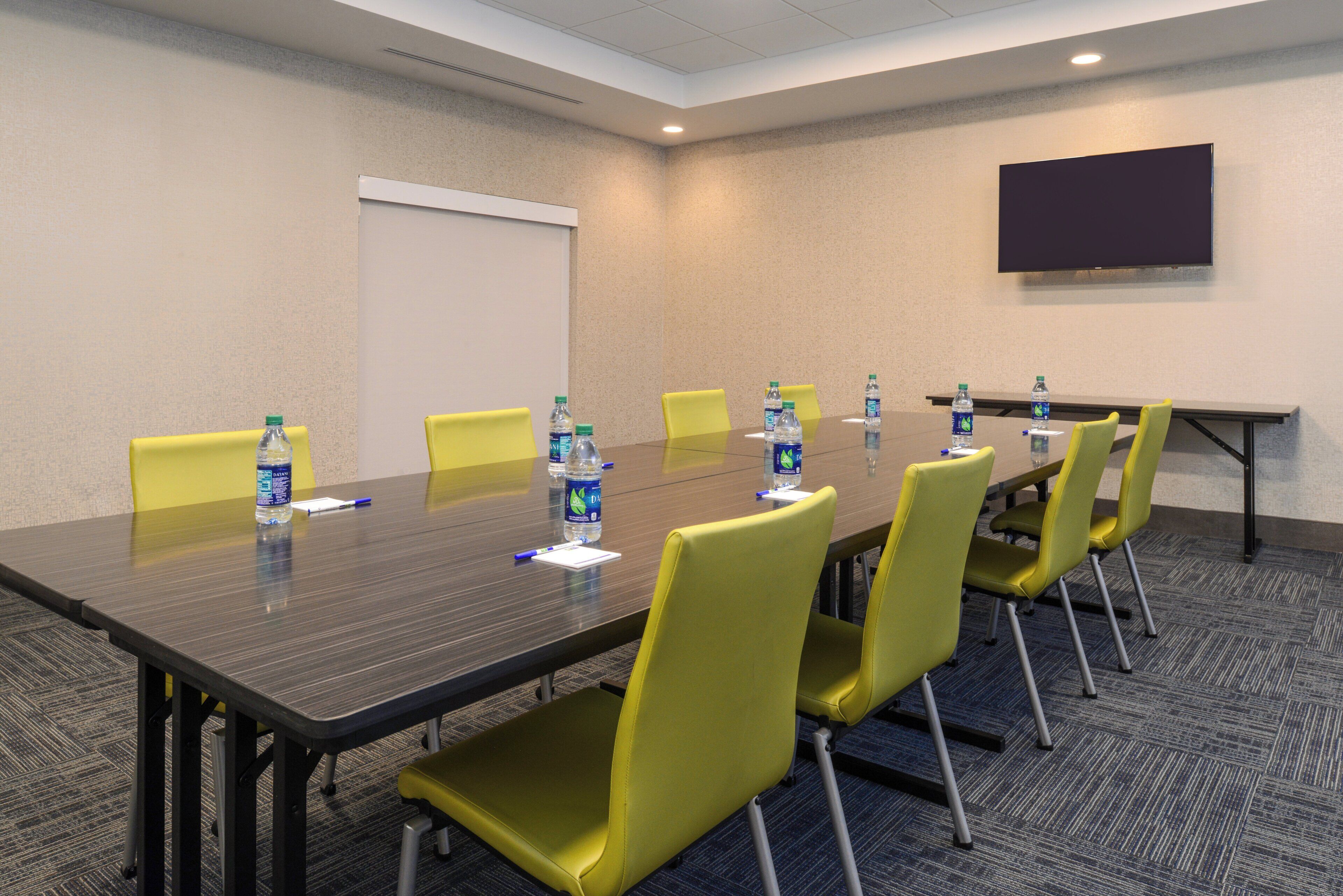 Un salón de conferencias con una mesa larga, sillas amarillas, botellas de agua y una pantalla de proyector en la pared.