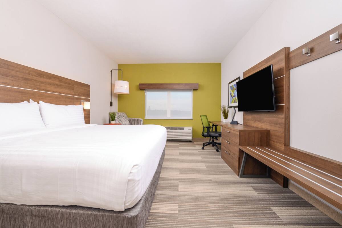 A hotel room with a large bed, a desk with a chair, a flat-screen TV, and a window with blinds.