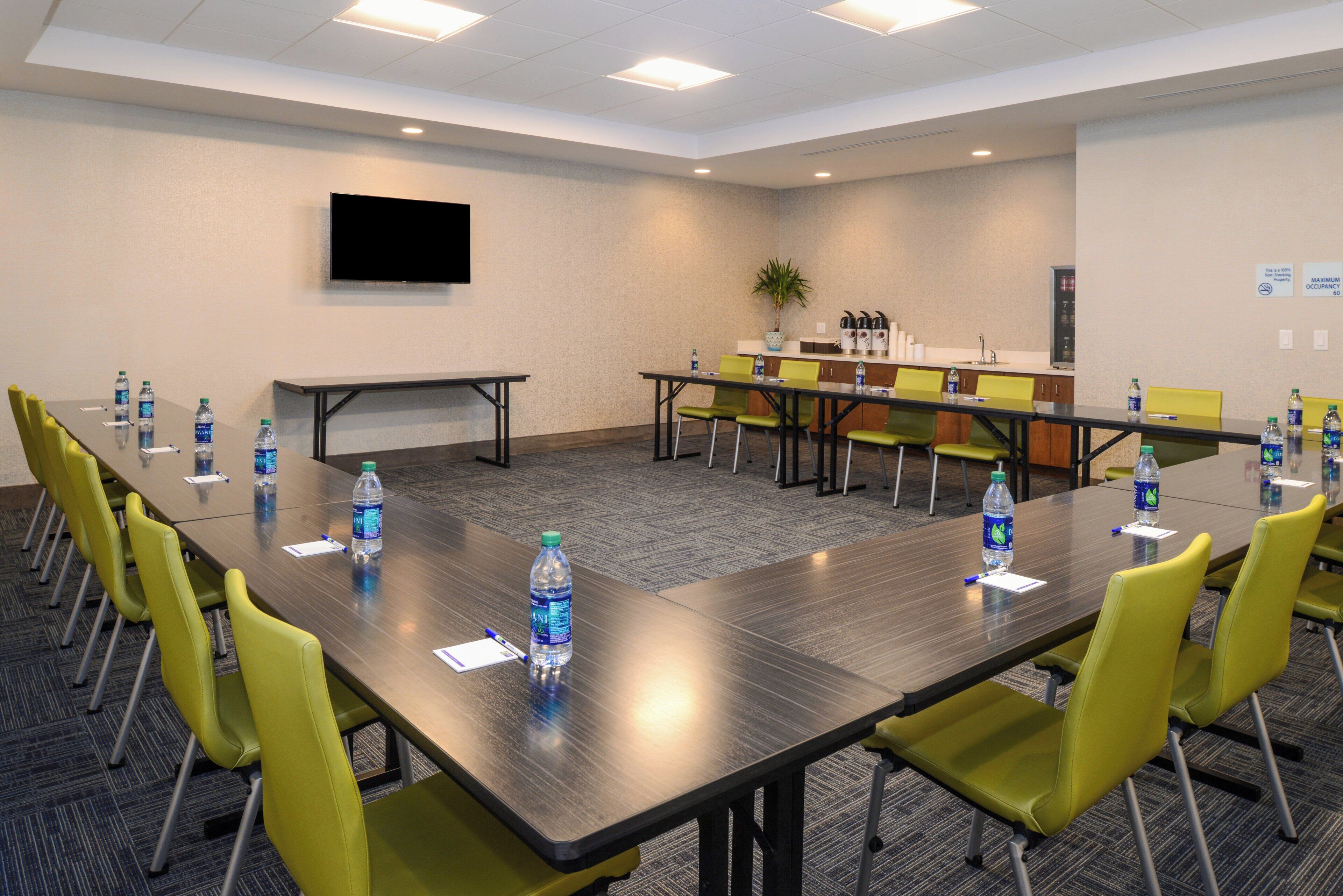 Un salón de conferencias con una larga mesa, sillas amarillas, botellas de agua, un televisor en la pared y una zona de kitchenette.