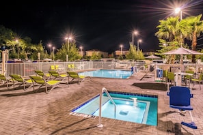 Un área de piscina nocturna con dos piscinas, sillas de descanso y sombrillas.