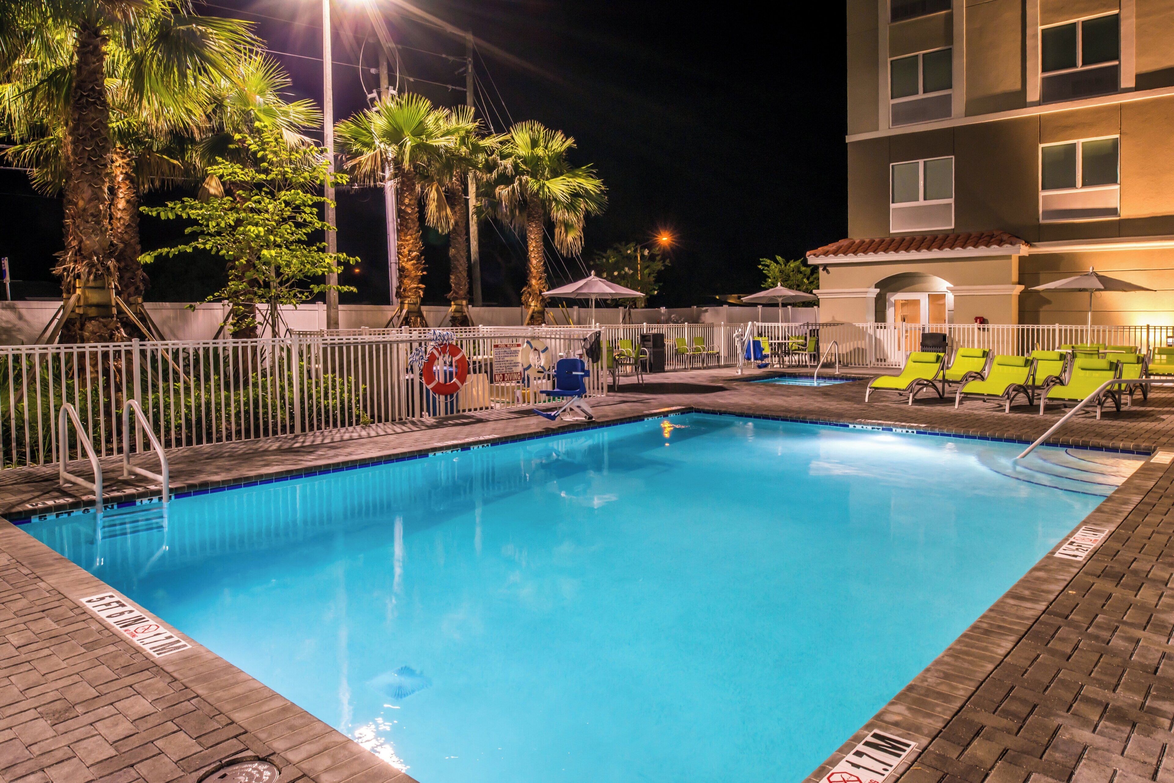 A nighttime swimming pool area with a fence, palm trees, and lounge chairs.