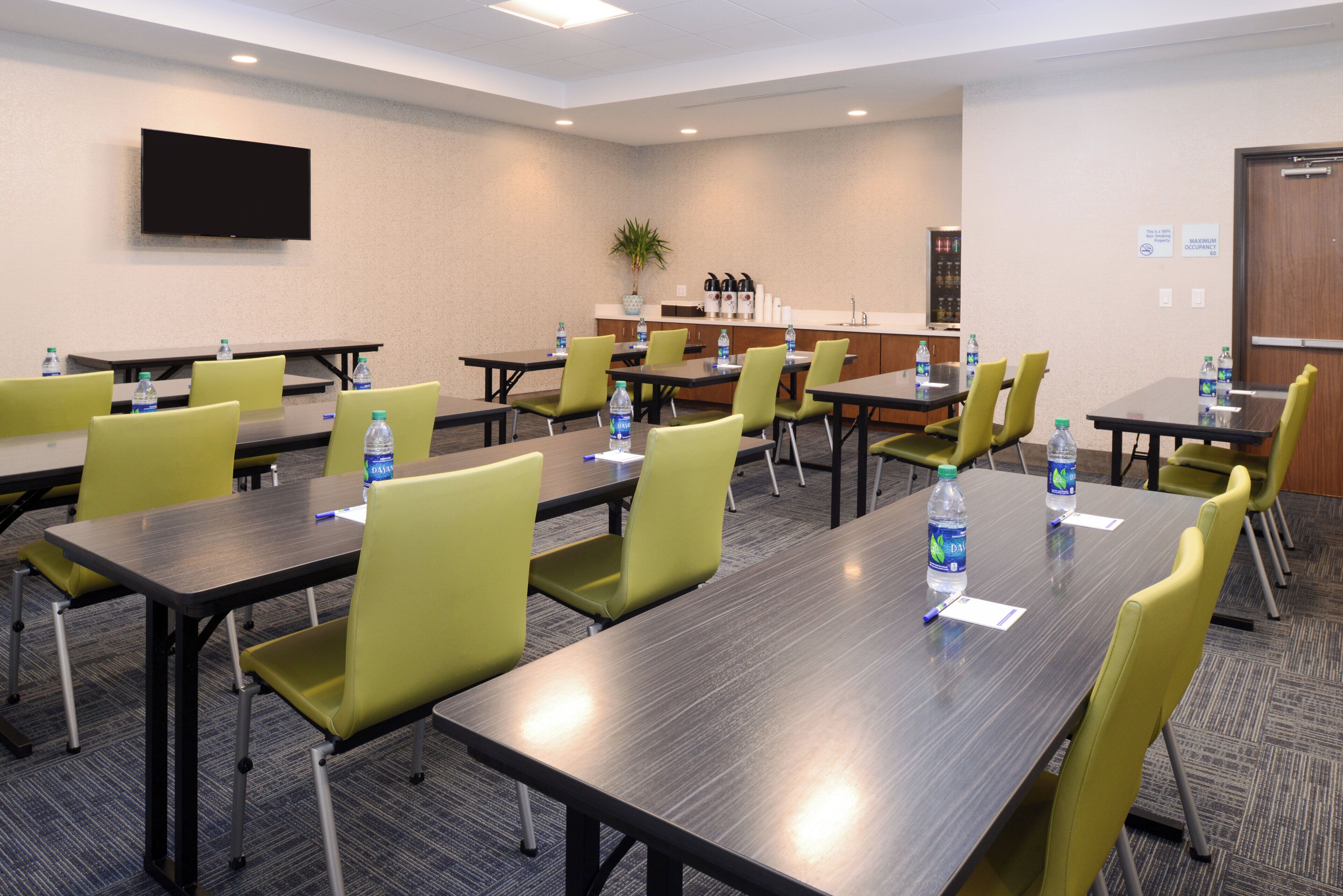 A conference room with multiple tables and chairs, water bottles on tables, a flat-screen TV on the wall, and a kitchenette area.