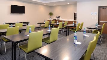 A conference room with multiple tables and chairs, water bottles on tables, a flat-screen TV on the wall, and a kitchenette area.