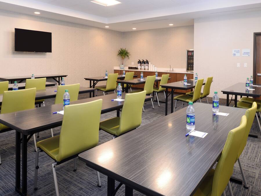 A conference room with multiple tables and chairs, water bottles on tables, a flat-screen TV on the wall, and a kitchenette area.