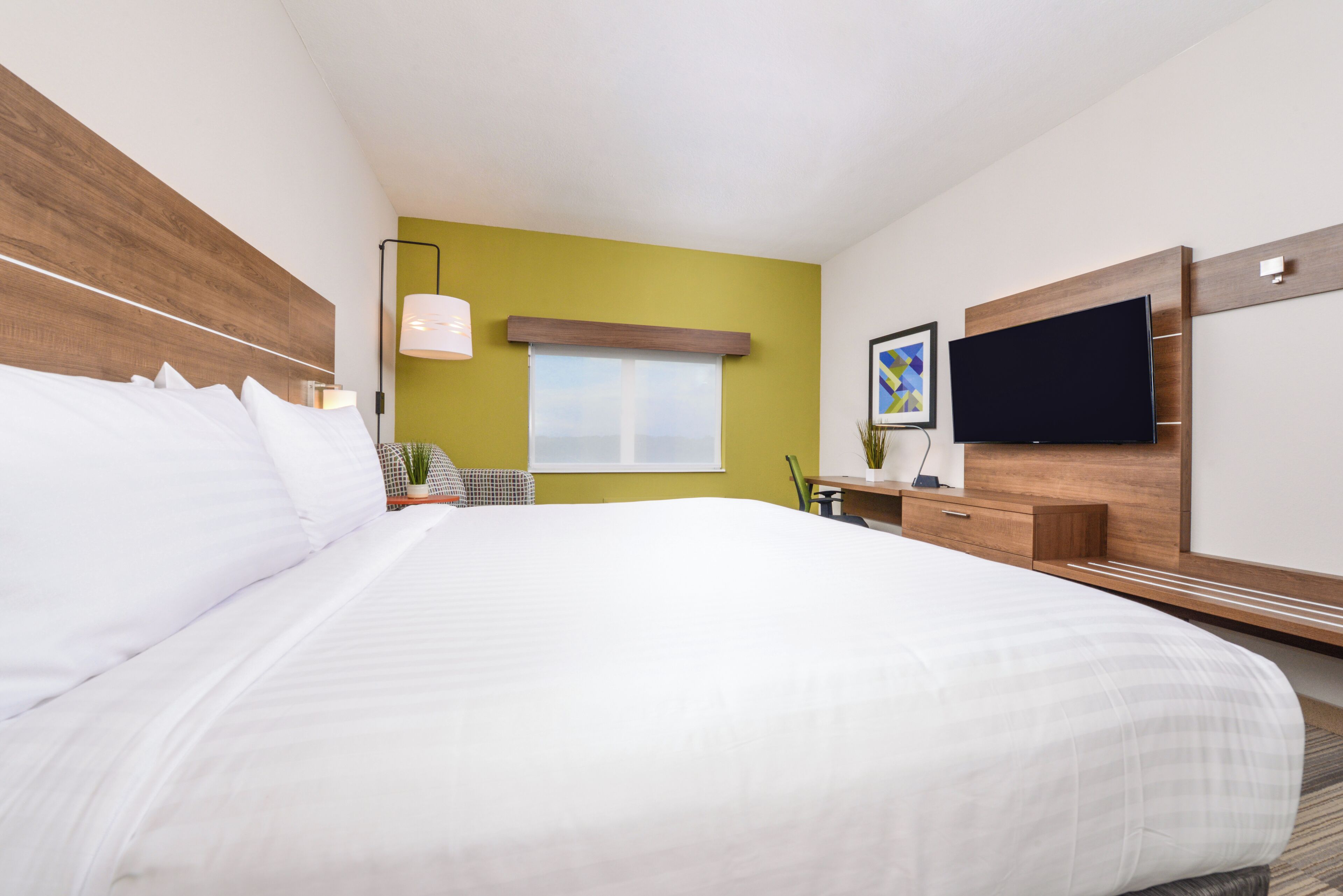A hotel room with a large bed, a flat-screen TV mounted on the wall, a window with a view, and a small table with a lamp.