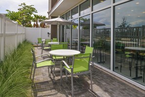 Un patio al aire libre con mesas redondas y sillas verdes, rodeado por una valla blanca y paredes de vidrio.