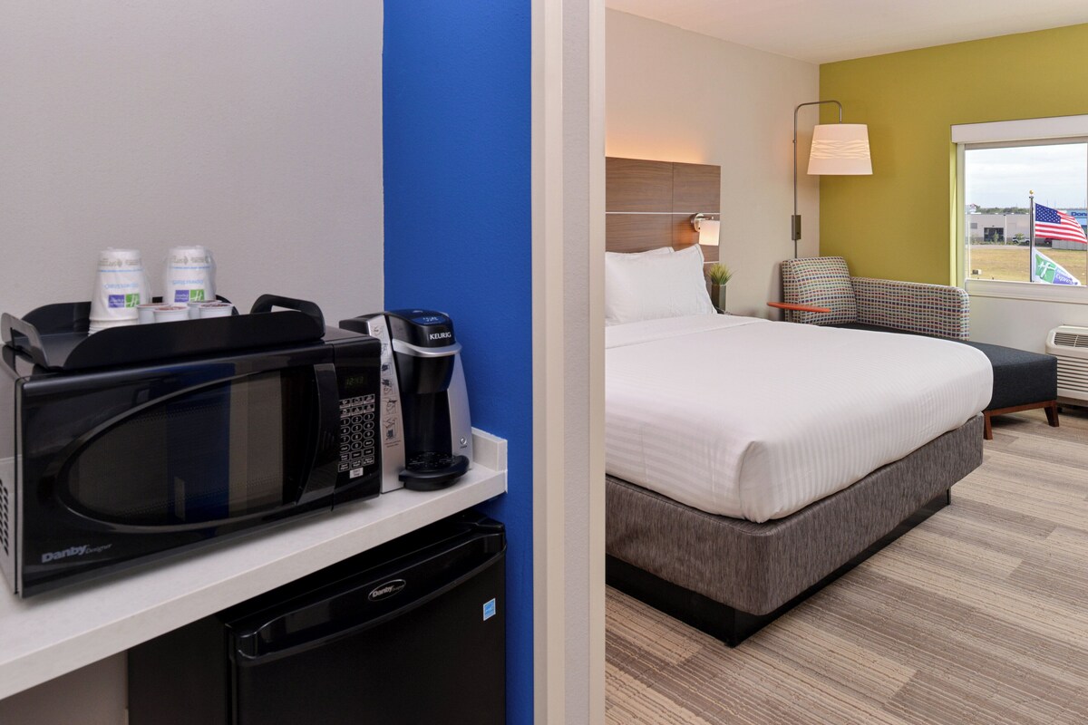 Habitación de hotel con cama, microondas, cafetera, refrigerador pequeño y ventana con la bandera estadounidense.