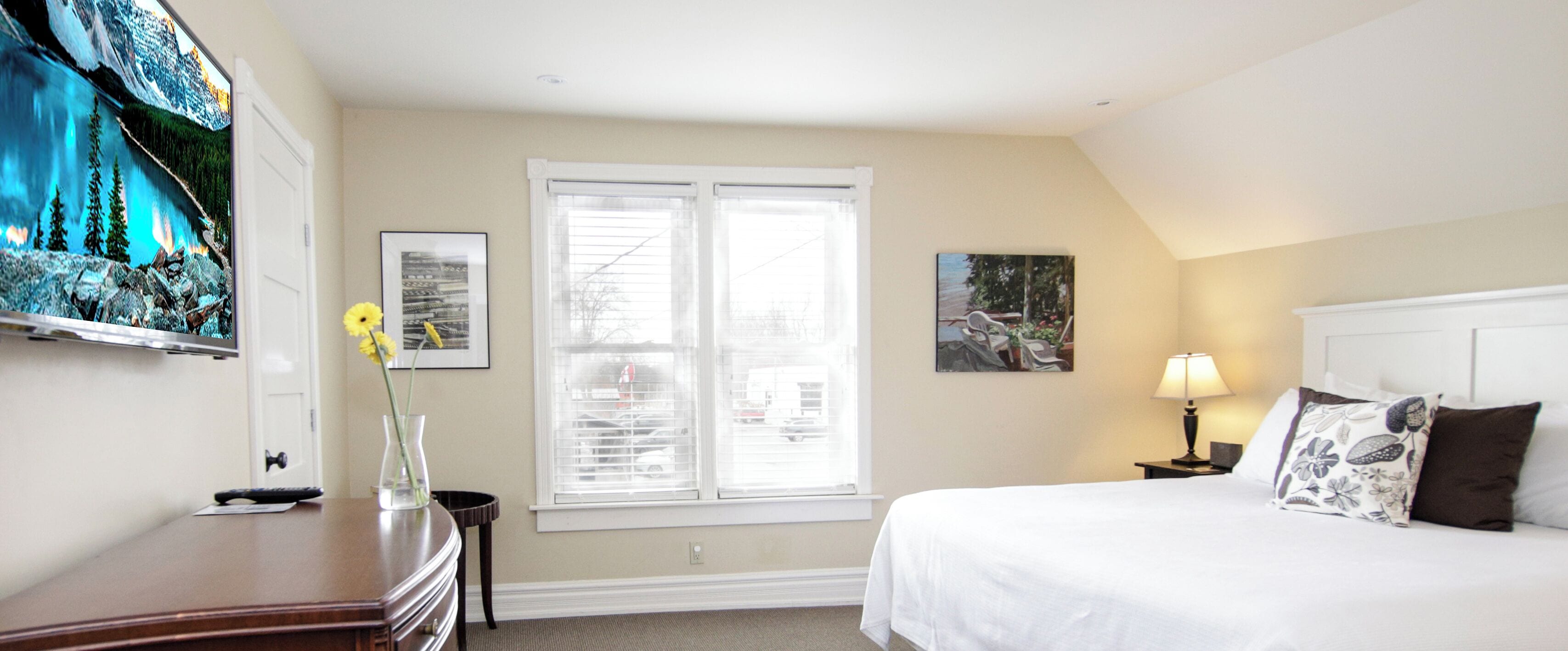 A hotel room with a bed, a desk, a vase with flowers, a window with blinds, and wall art.
