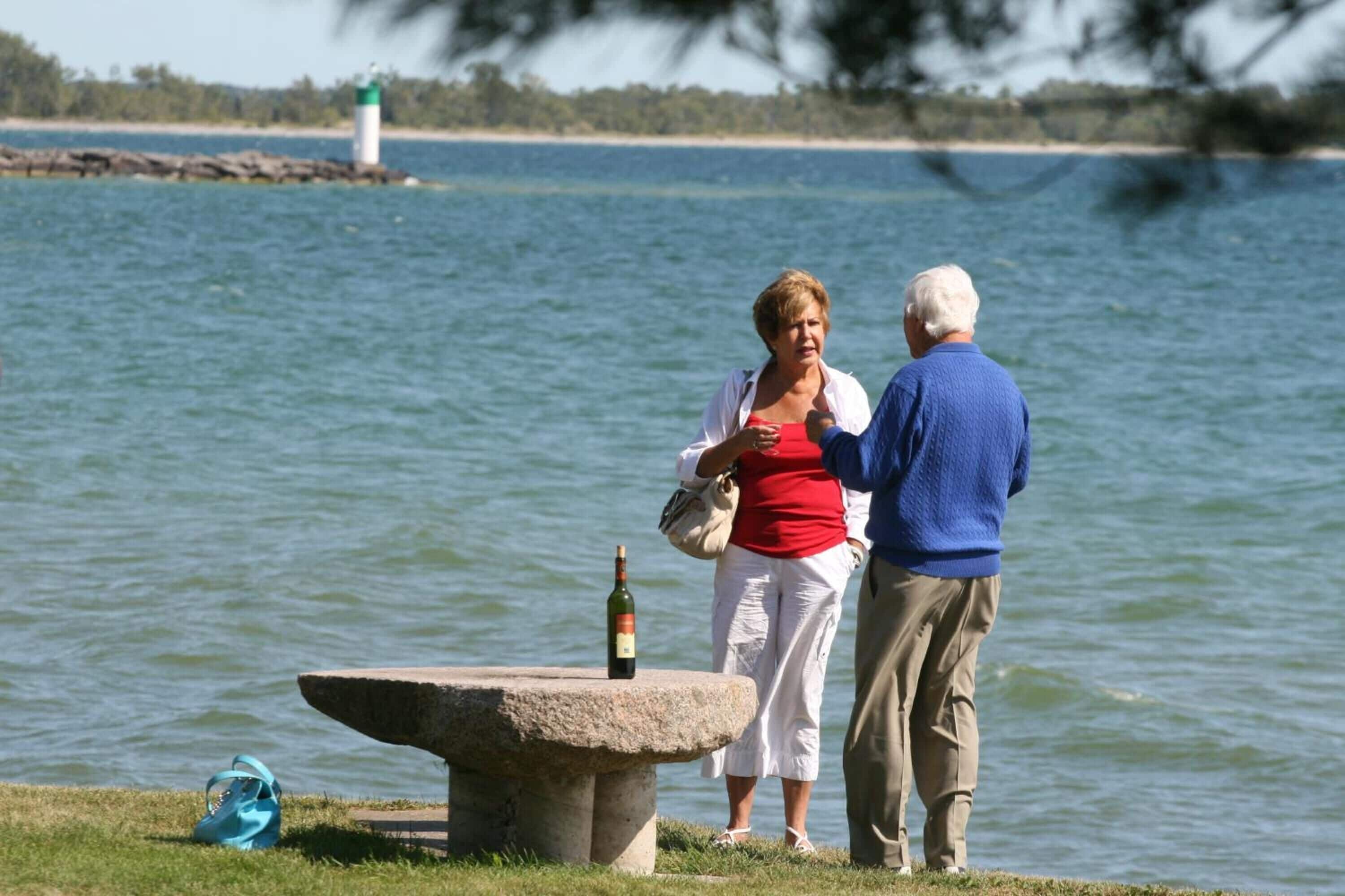 Two people standing by a stone bench with a bottle on it, overlooking a body of water with a lighthouse in the distance.
