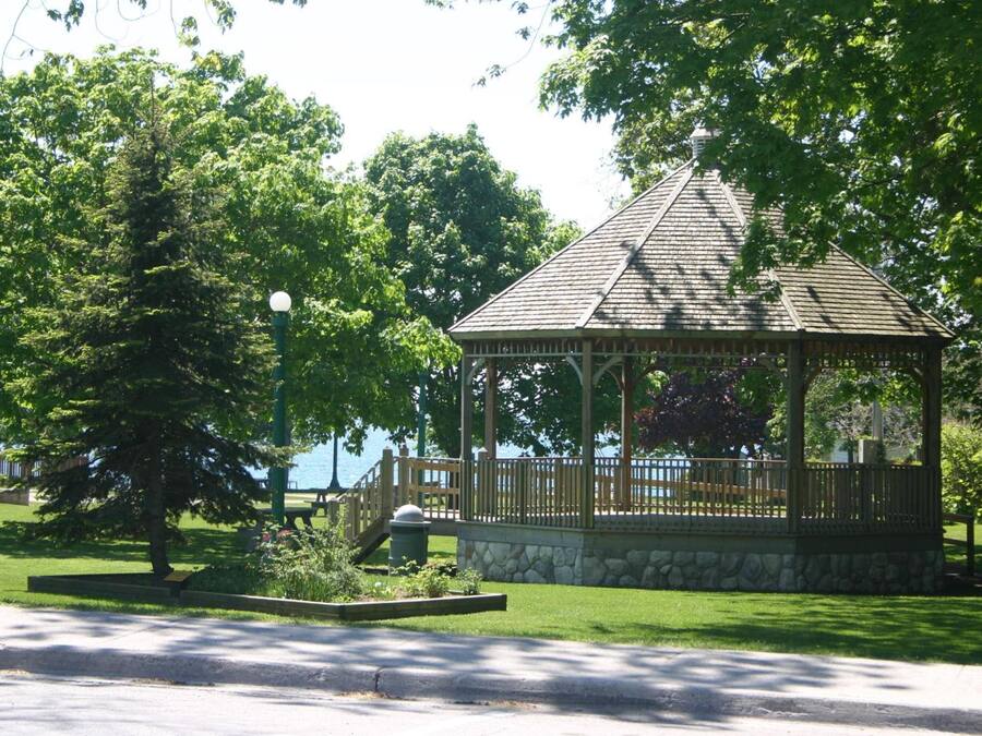 A gazebo with a stone base and wooden structure, surrounded by a grassy area and trees.