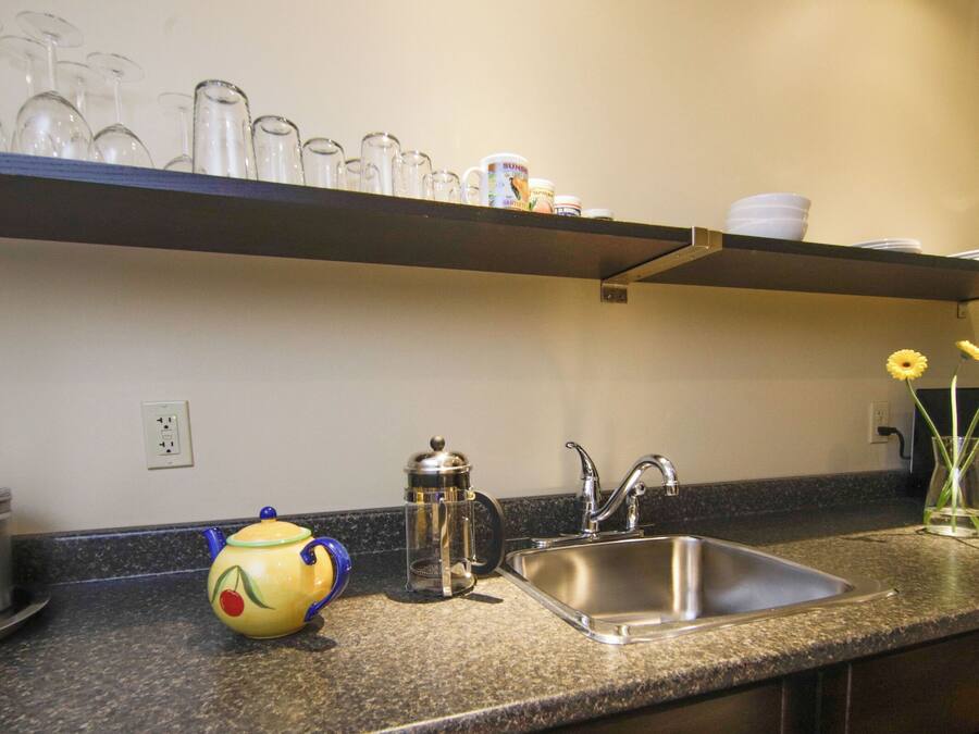 A kitchen with a stainless steel sink, a coffee maker, and a shelf above the counter holding glassware and a mug.