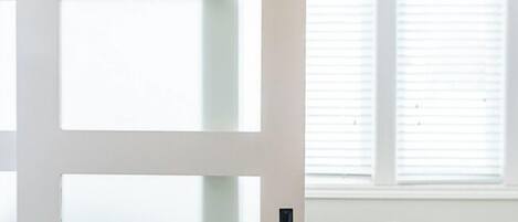 A room with a frosted glass door, a window with blinds, a white bean bag, and a wooden table.