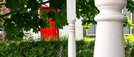 Un jardín con una estructura de columna blanca, un edificio rojo al fondo y diversas plantas y flores.