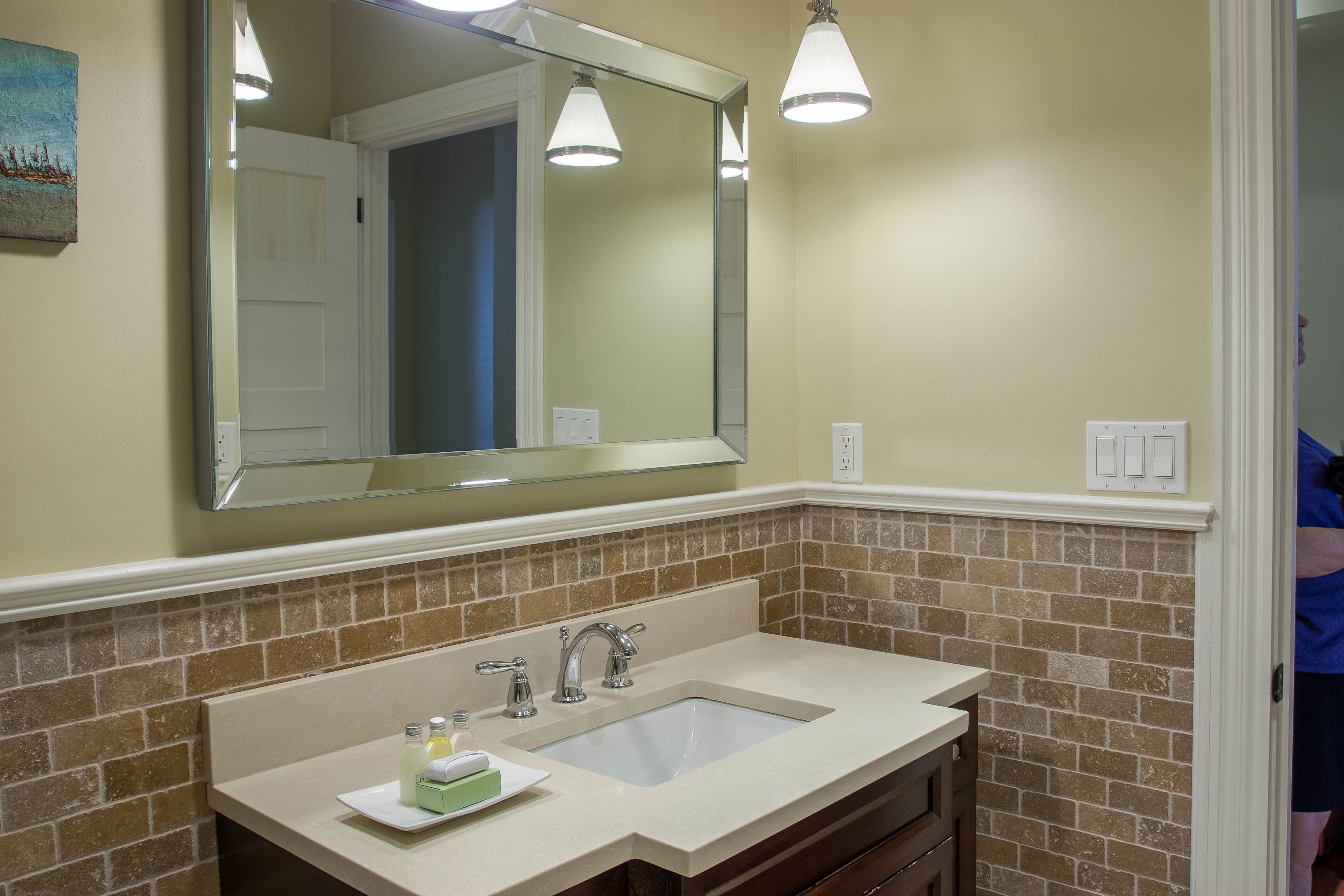 A bathroom with a beige sink, two faucets, a large mirror, and a light fixture above.