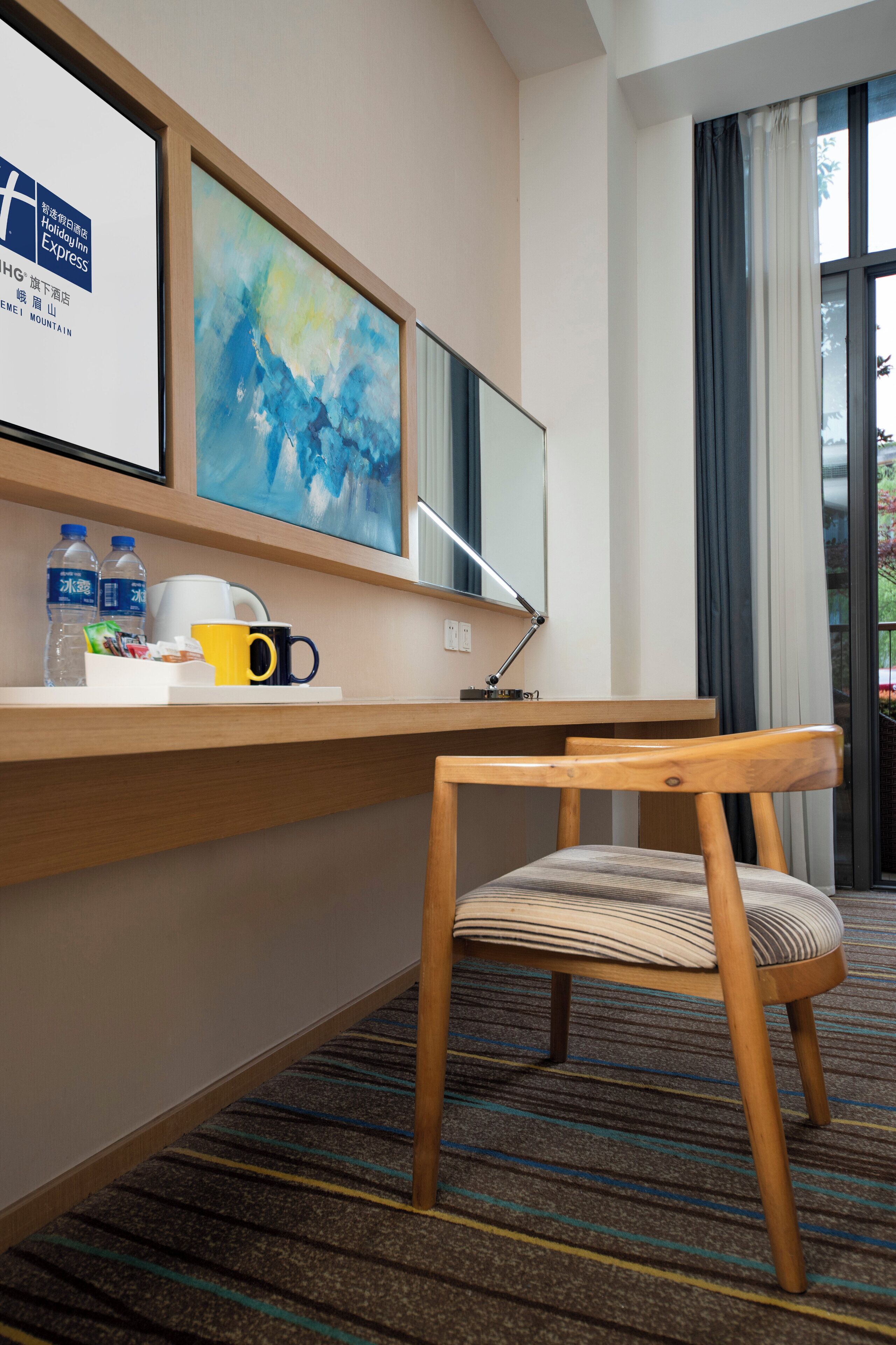 A hotel room with a wooden desk, a chair, a framed artwork, a water bottle, a mug, and a window with curtains.