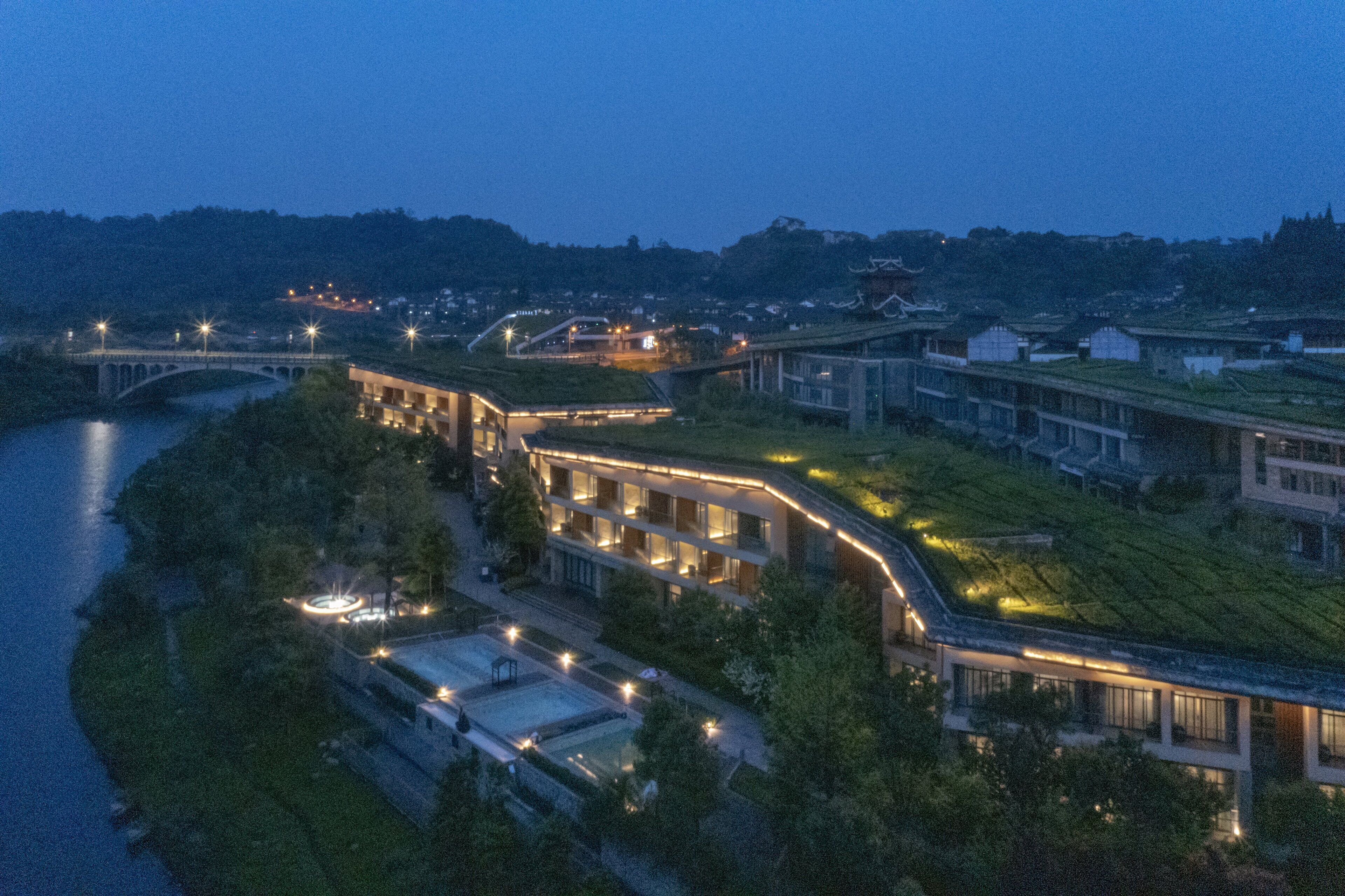 A modern hotel with a green roof, a river in the foreground, and a bridge in the background.