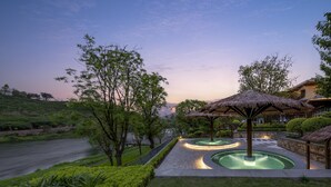 A landscaped garden with a pool, thatched-roof structures, and a view of a distant hill. - Holiday Inn Express Emei Mountain by IHG (Leshan)