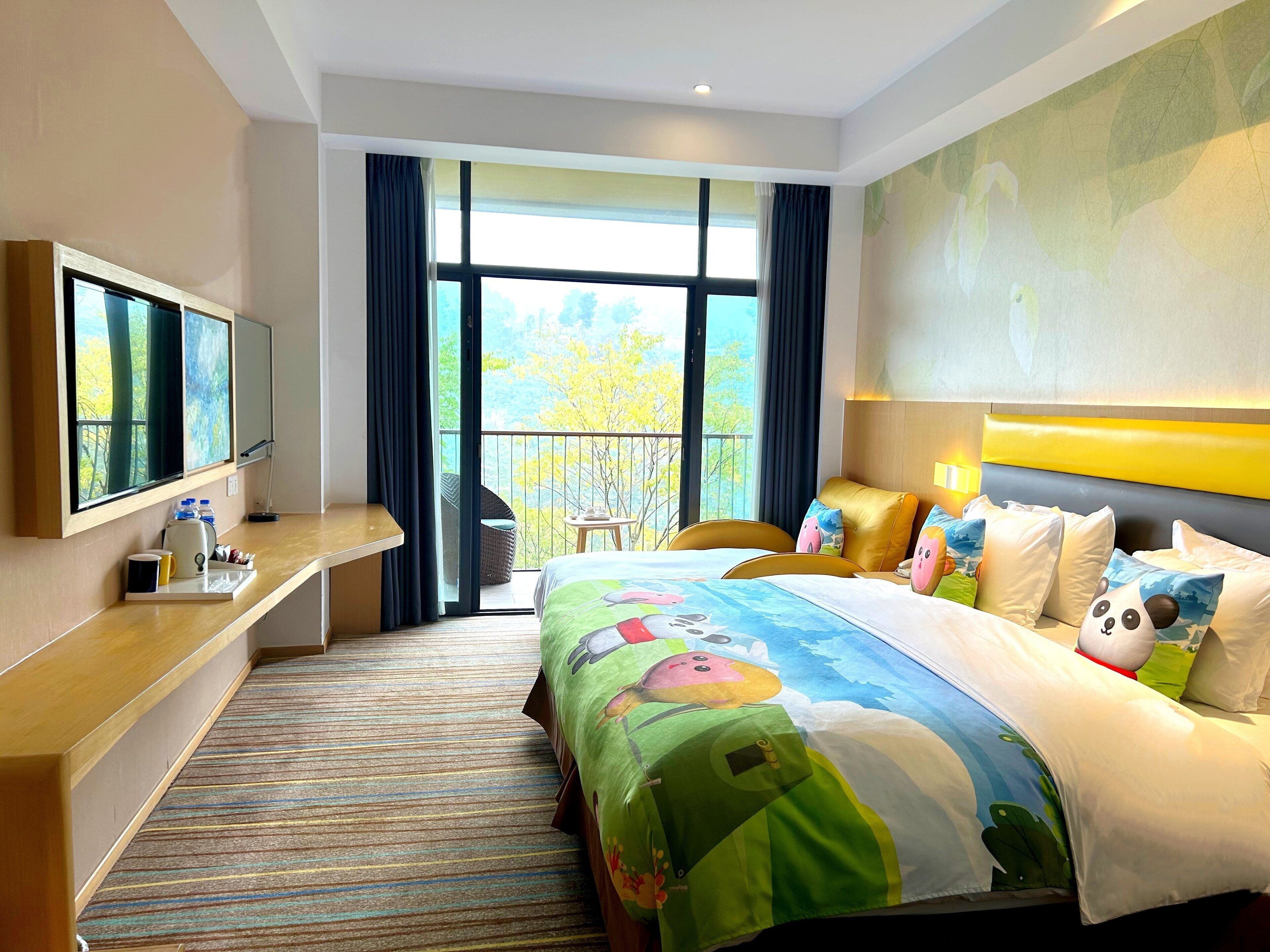 A hotel room with a large bed, a desk with a flat-screen TV, a balcony with a chair, and a view of trees.