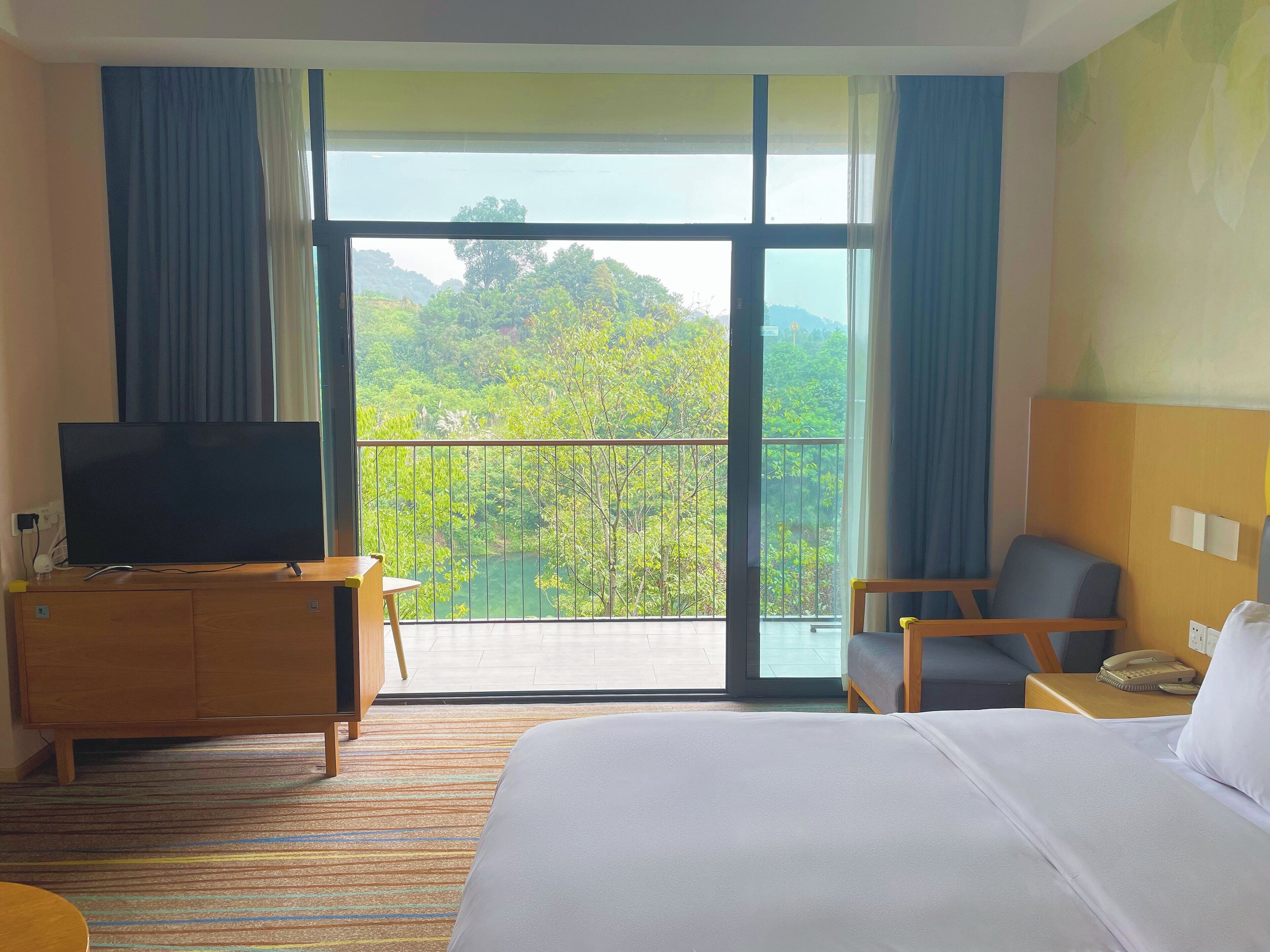 A hotel room with a large bed, a TV on a wooden cabinet, a chair, a small table, and a balcony with a view of greenery.
