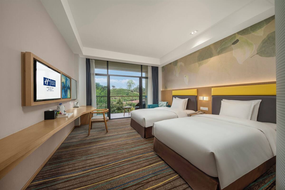 A hotel room with two beds, a desk, a chair, a television, and a balcony with a view of greenery.