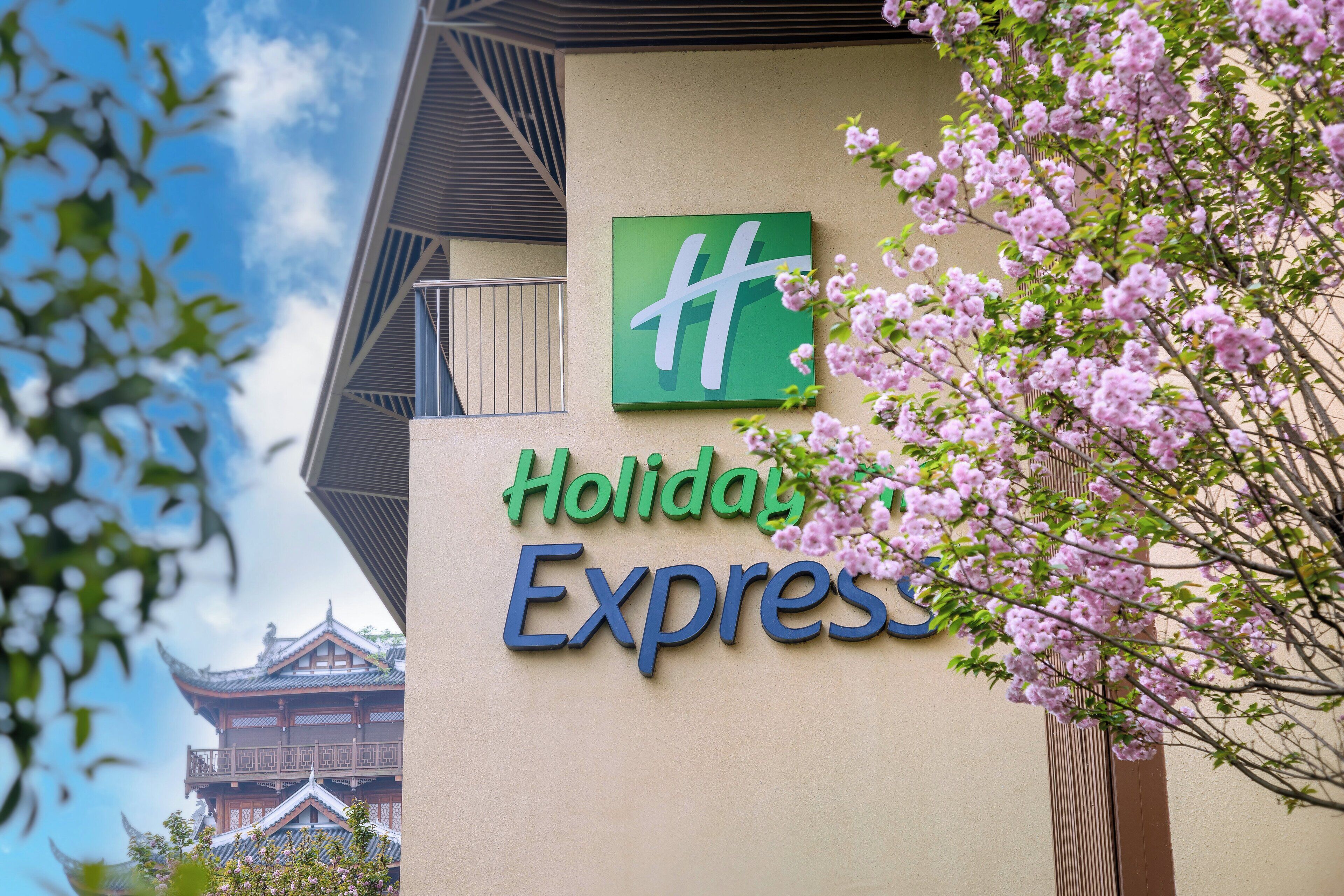 Un letrero de hotel con el logotipo de Holiday Inn Express, un edificio tradicional al fondo y flores de cerezo en primer plano.