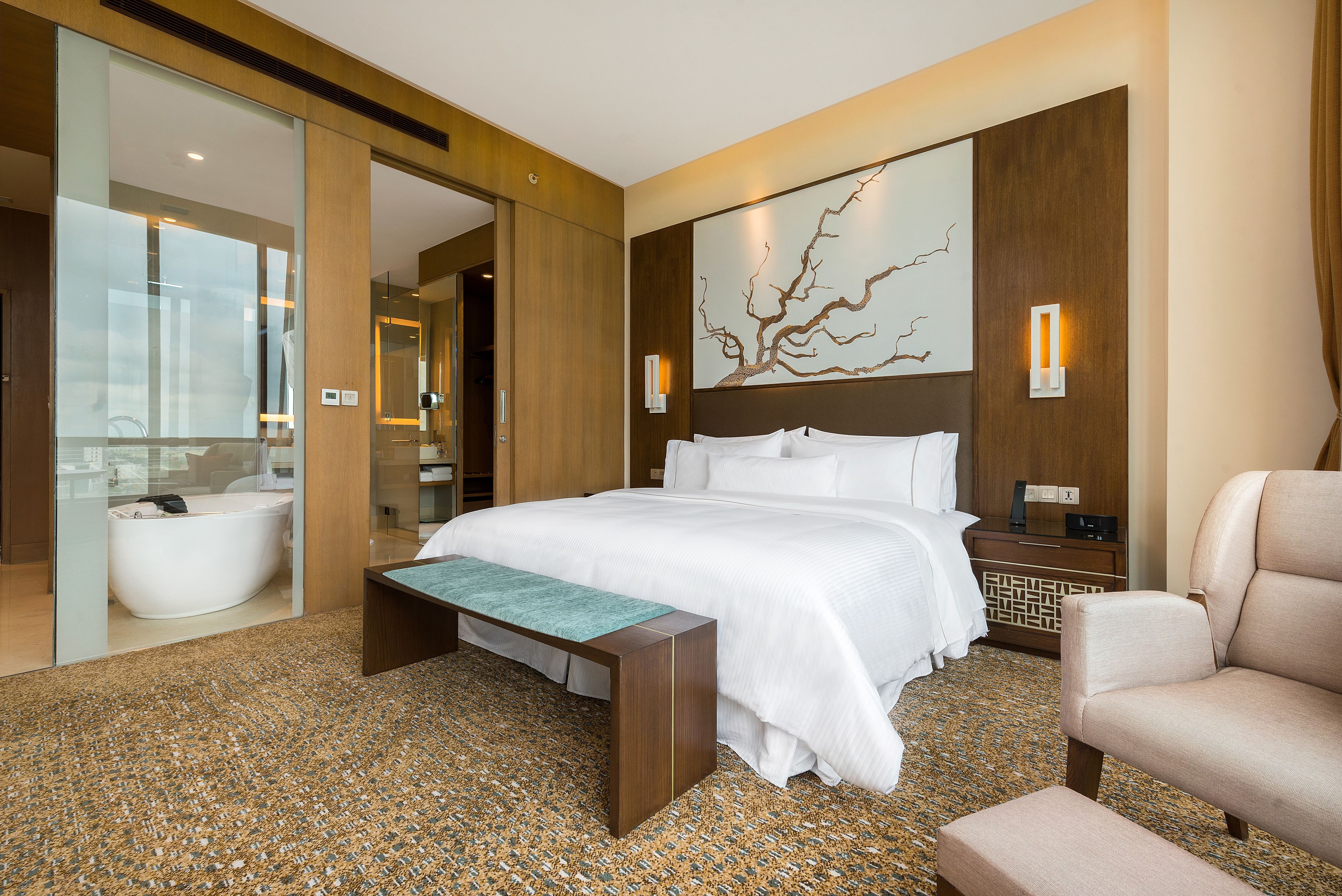 Habitación de hotel moderna con una cama grande, un banco, una silla, una bañera y una obra de arte colgada en la pared.