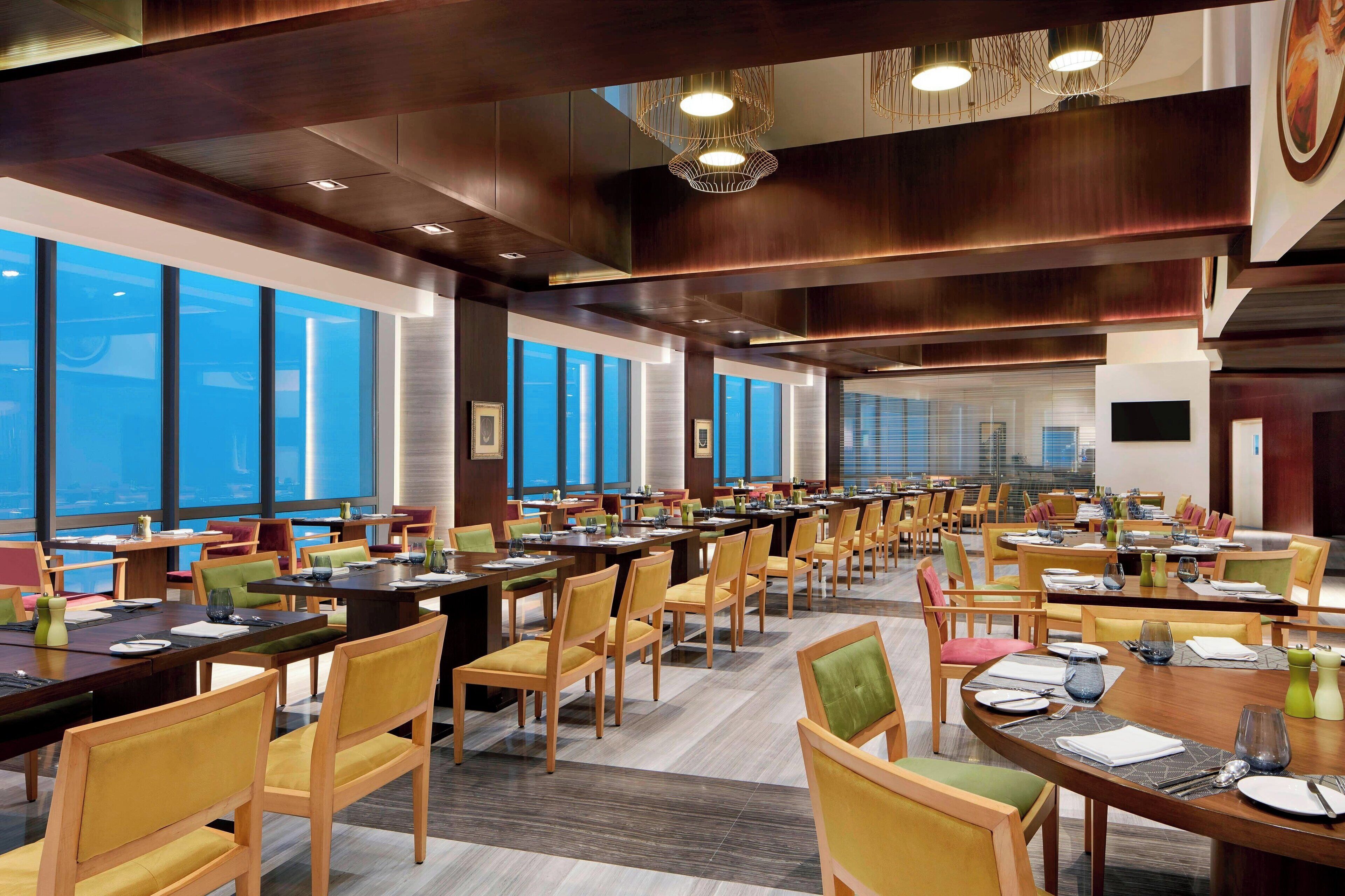 A spacious dining area with multiple tables and chairs, large windows offering views, and modern lighting fixtures.
