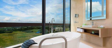 A modern bathroom with a large bathtub, a vanity with a mirror, and a view of a green landscape through large windows.