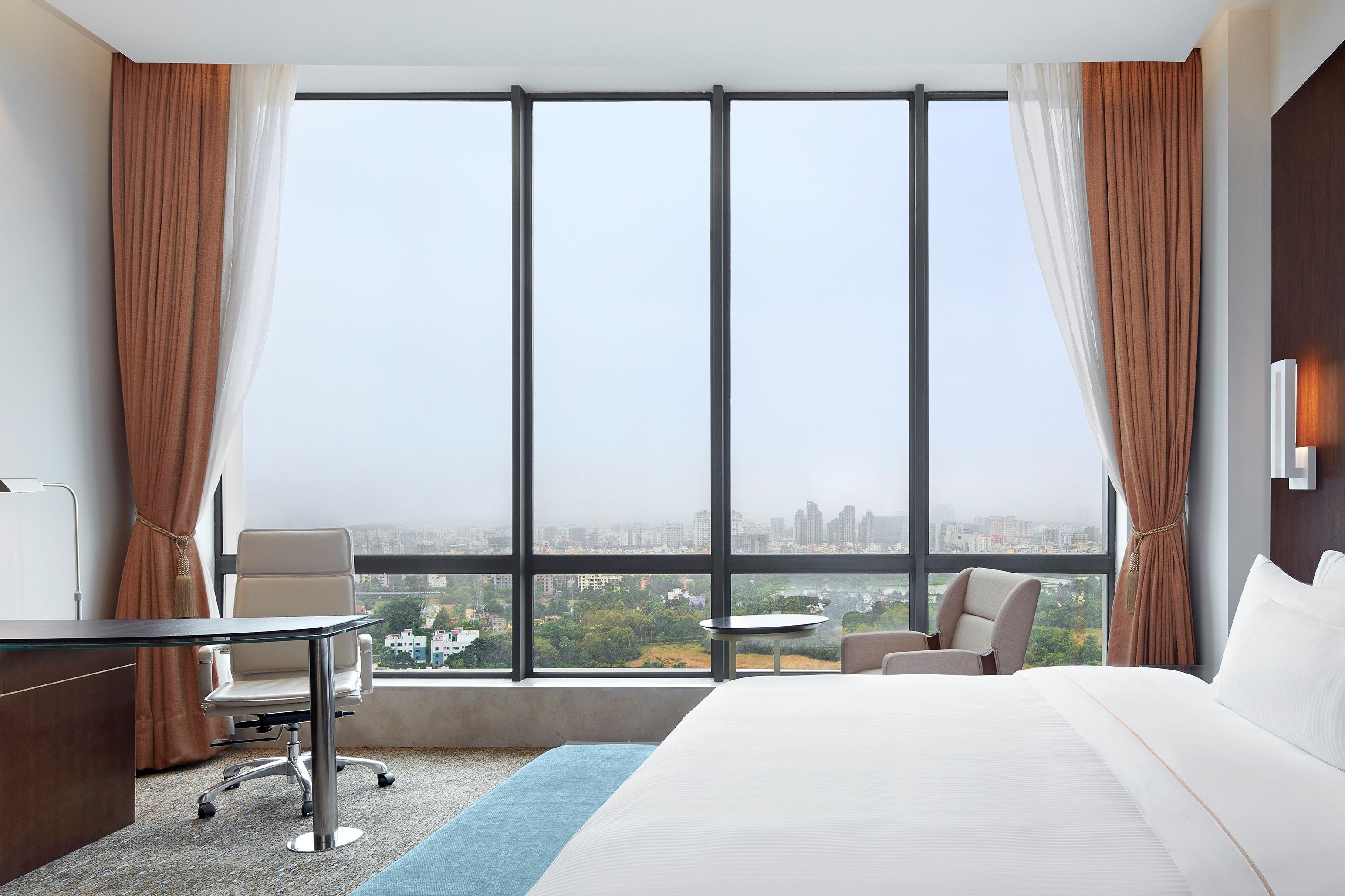 A hotel room with a large window, a bed, a desk, and two chairs.