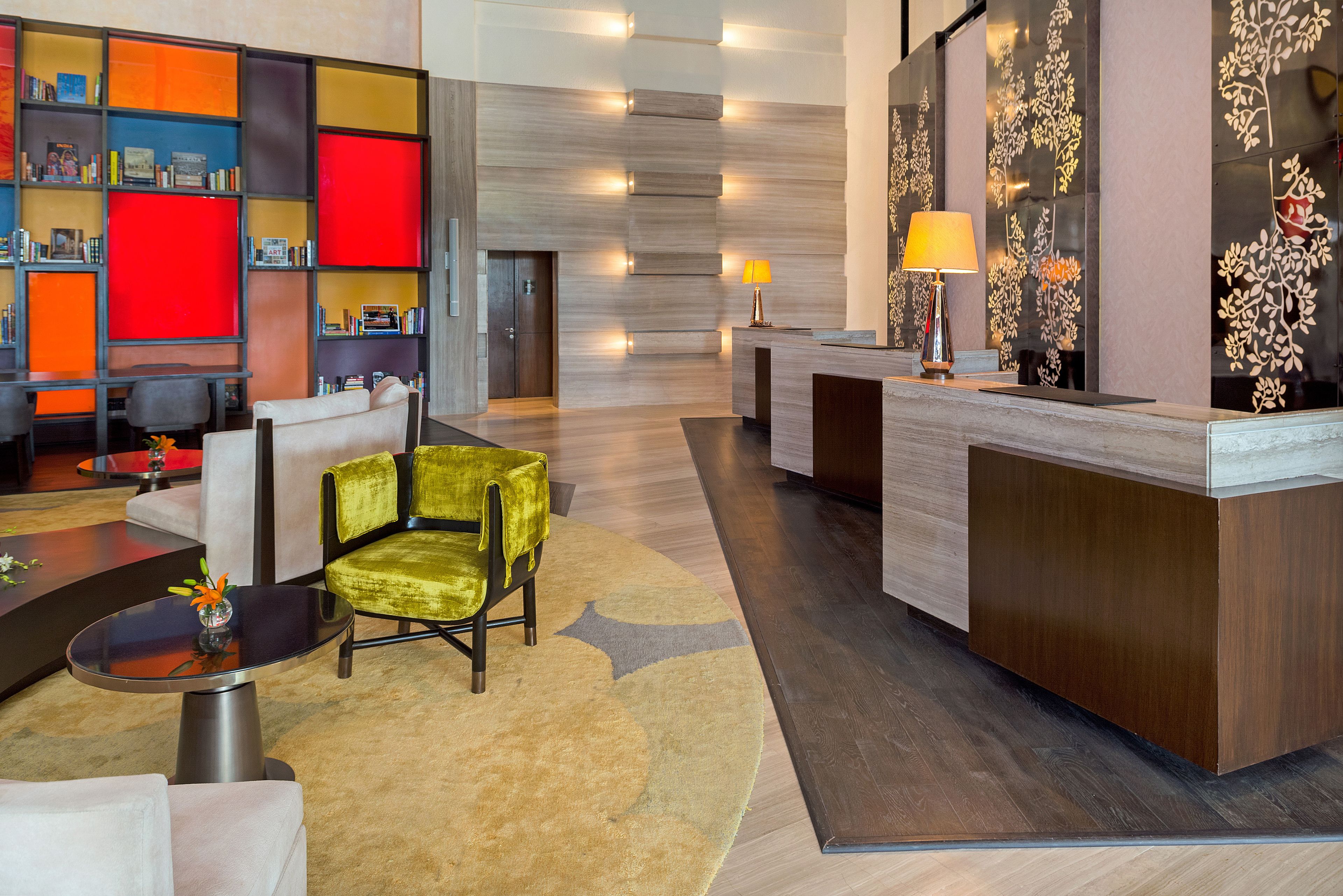 A hotel lobby with a reception desk, a seating area with a green chair, and a bookshelf with colorful compartments.