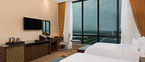 A hotel room with two beds, a desk with a chair, a TV, and a large window with curtains.