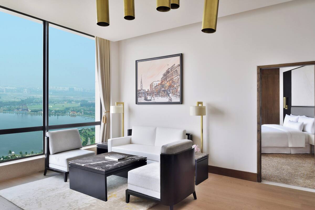 A modern hotel room with a large window offering a city view, a white sofa, a small table, and a separate bed area.