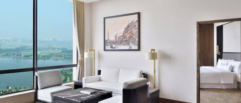 A modern hotel room with a large window offering a city view, a white sofa, a small table, and a separate bed area.