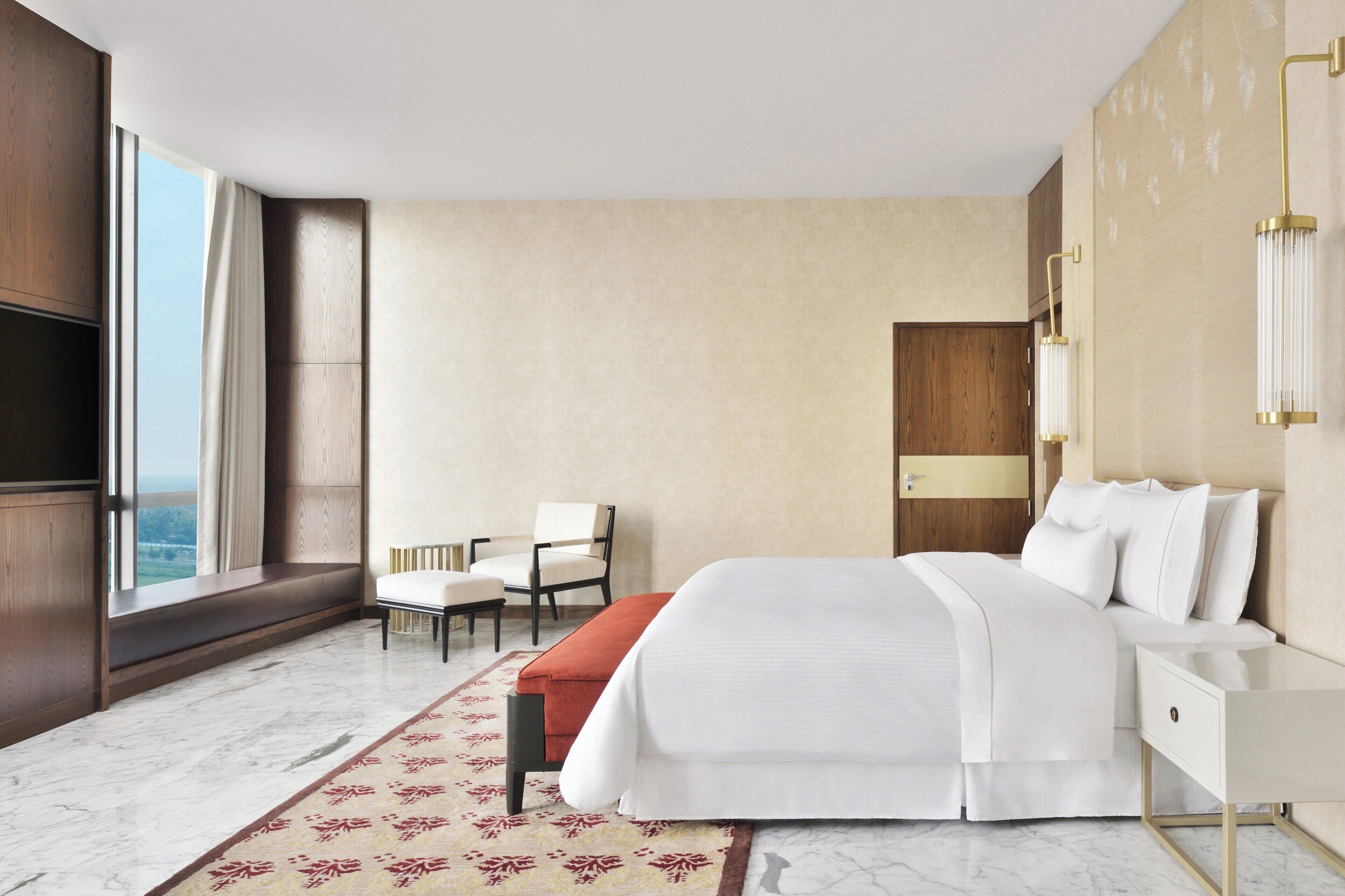 A modern hotel room with a large bed, a seating area with a chair, a desk, and a large window offering a city view.
