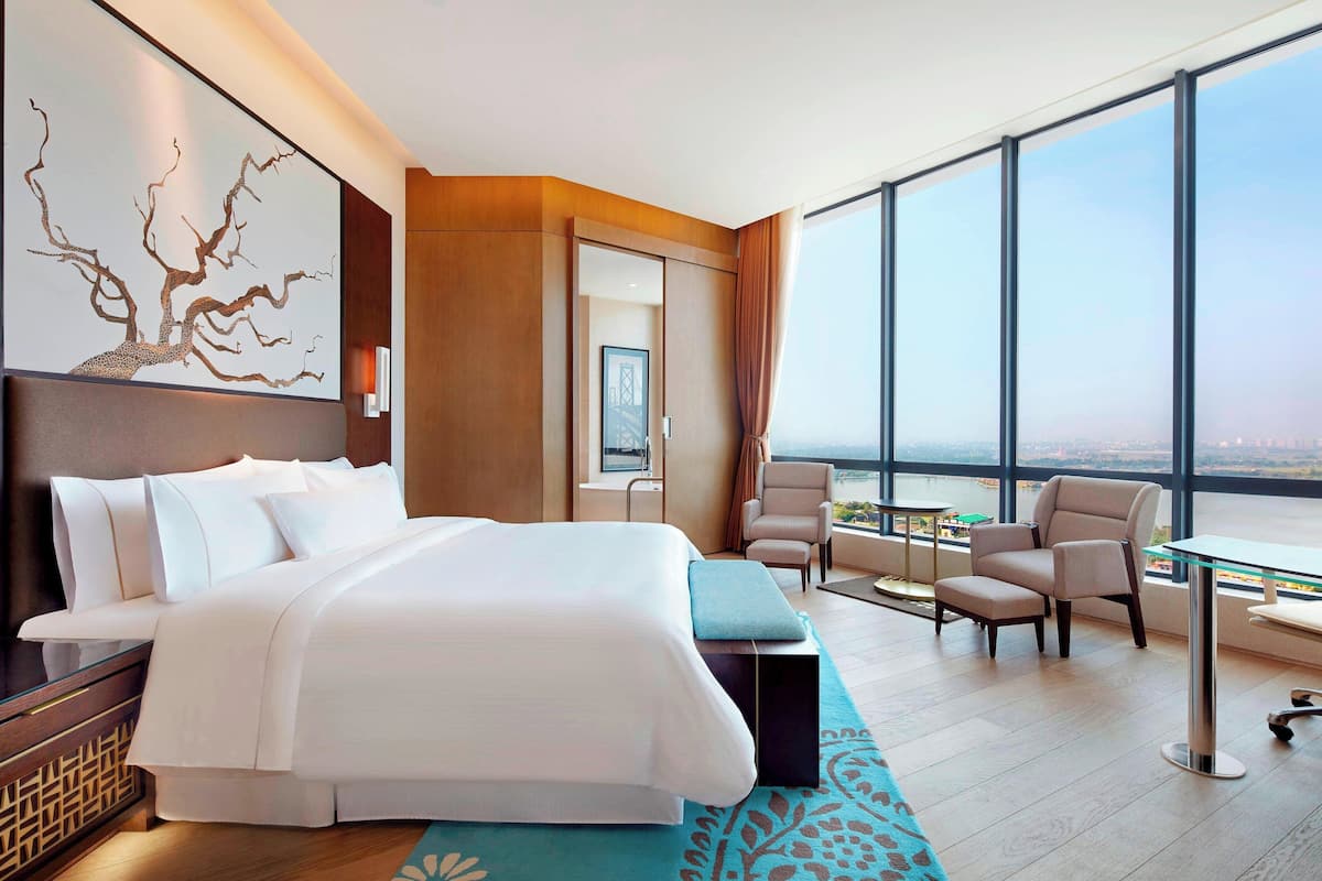 A modern hotel room with a large bed, a seating area with chairs, a table, and a view of the city through large windows.