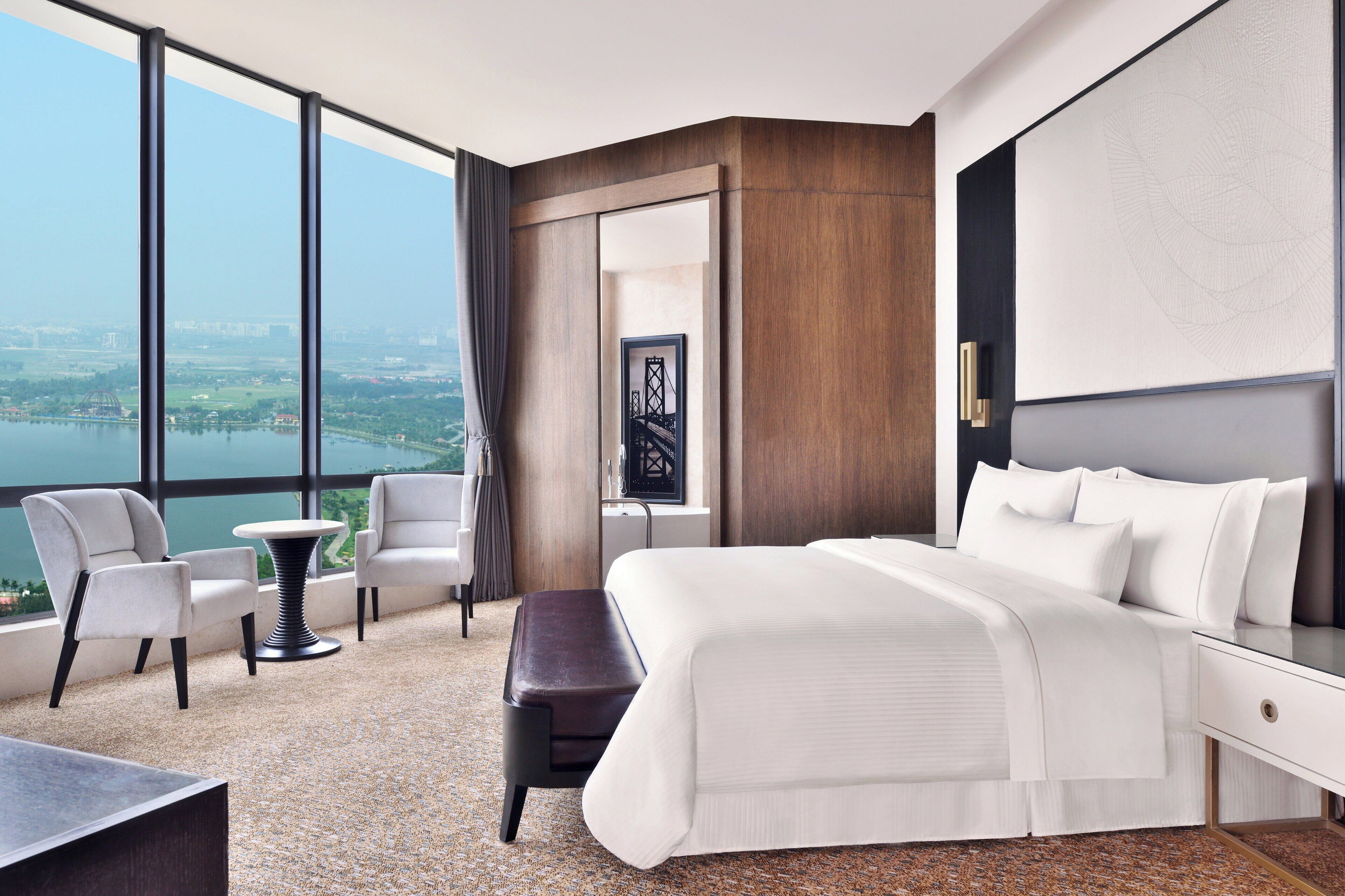 A modern hotel room with a large bed, a sitting area with two chairs, a small table, and a view of a cityscape and body of water through large windows.
