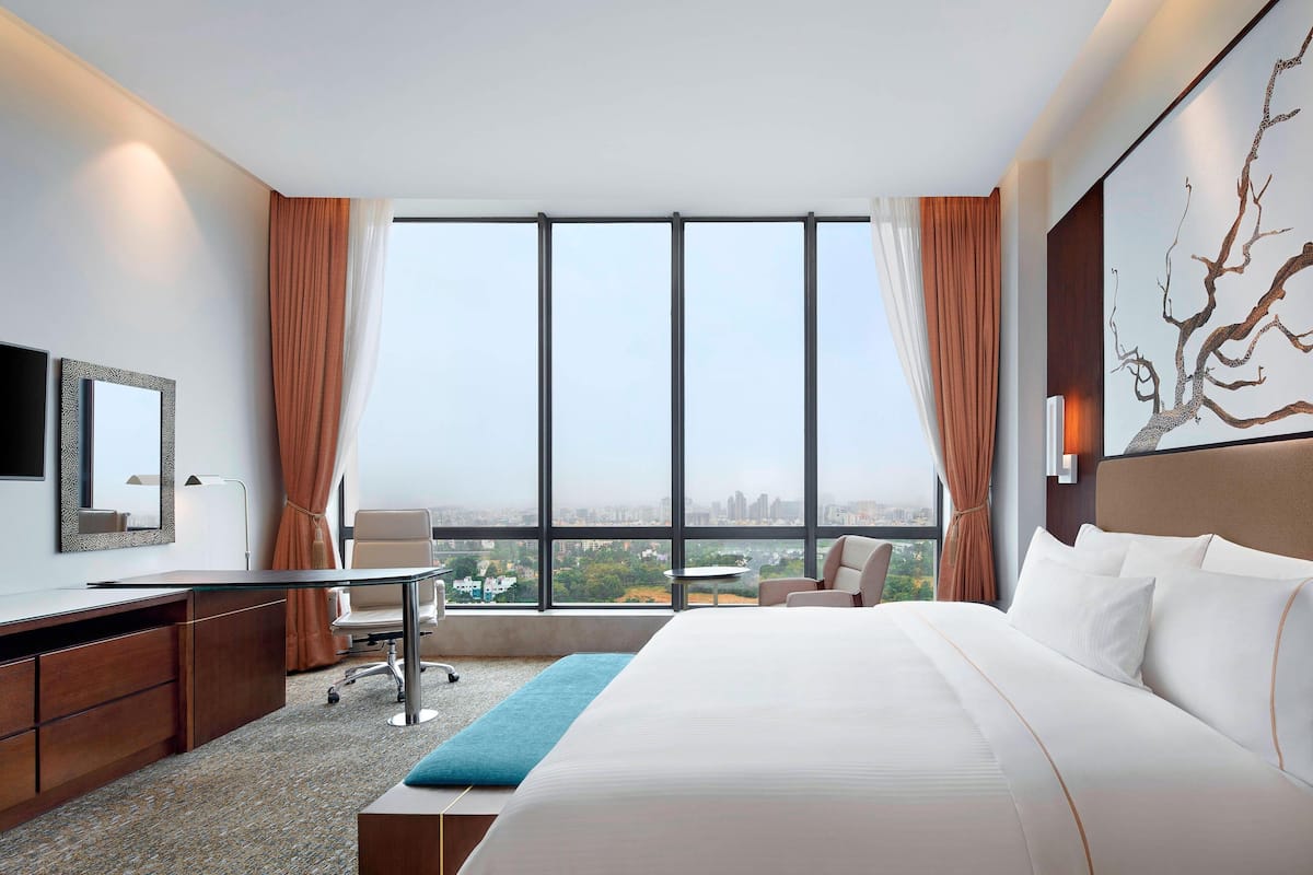 A hotel room with a large bed, a desk, a chair, and a view of the city through large windows.