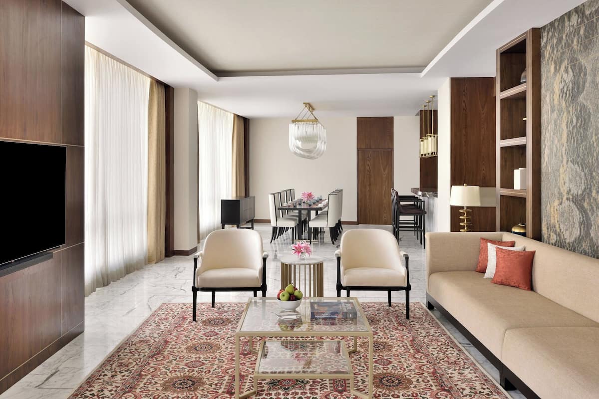 A modern living room with a sofa, two armchairs, a coffee table with fruit, a TV, and a dining area in the background.