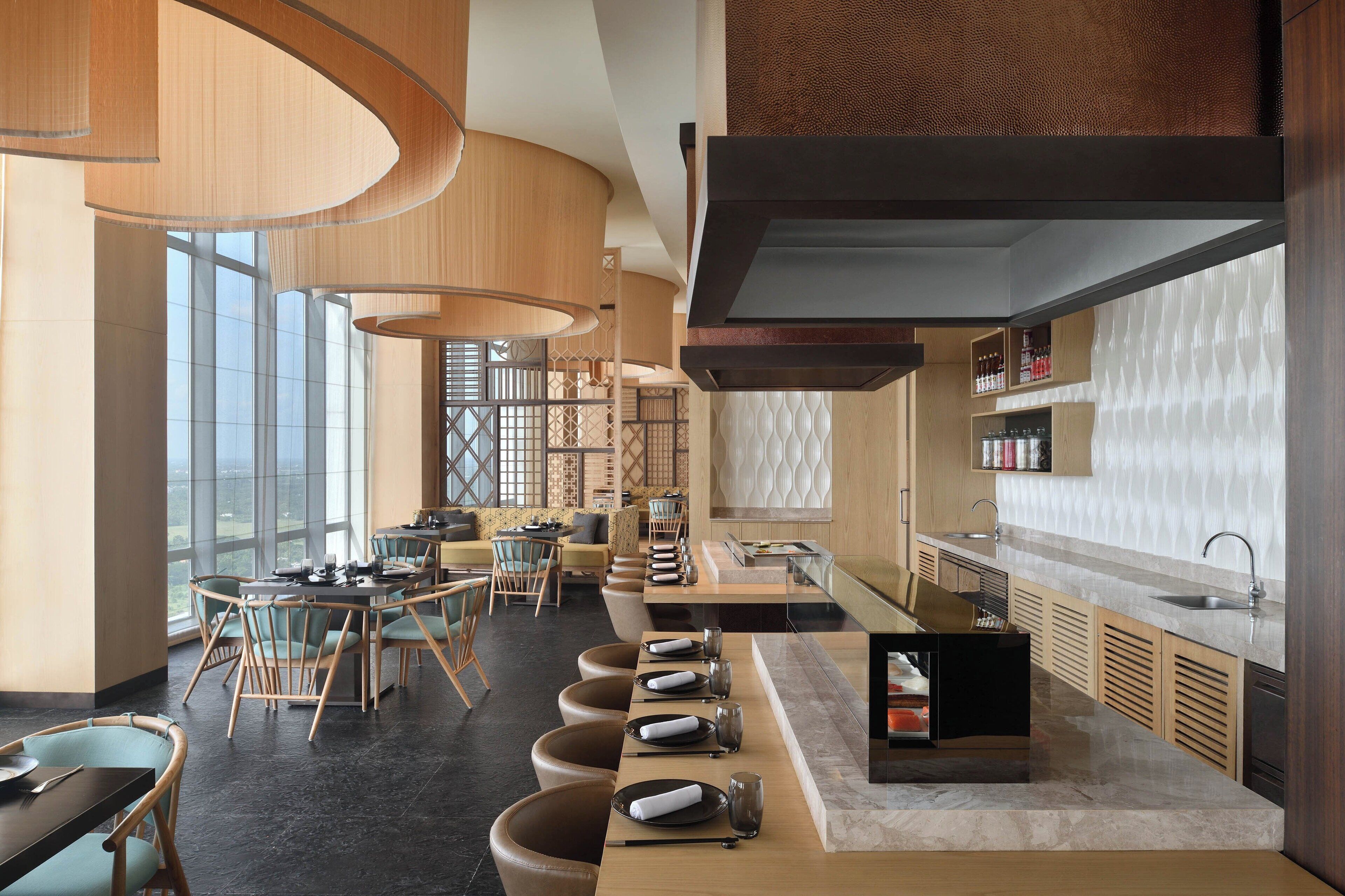 A modern restaurant with a long counter, bar seating, and a kitchen area.