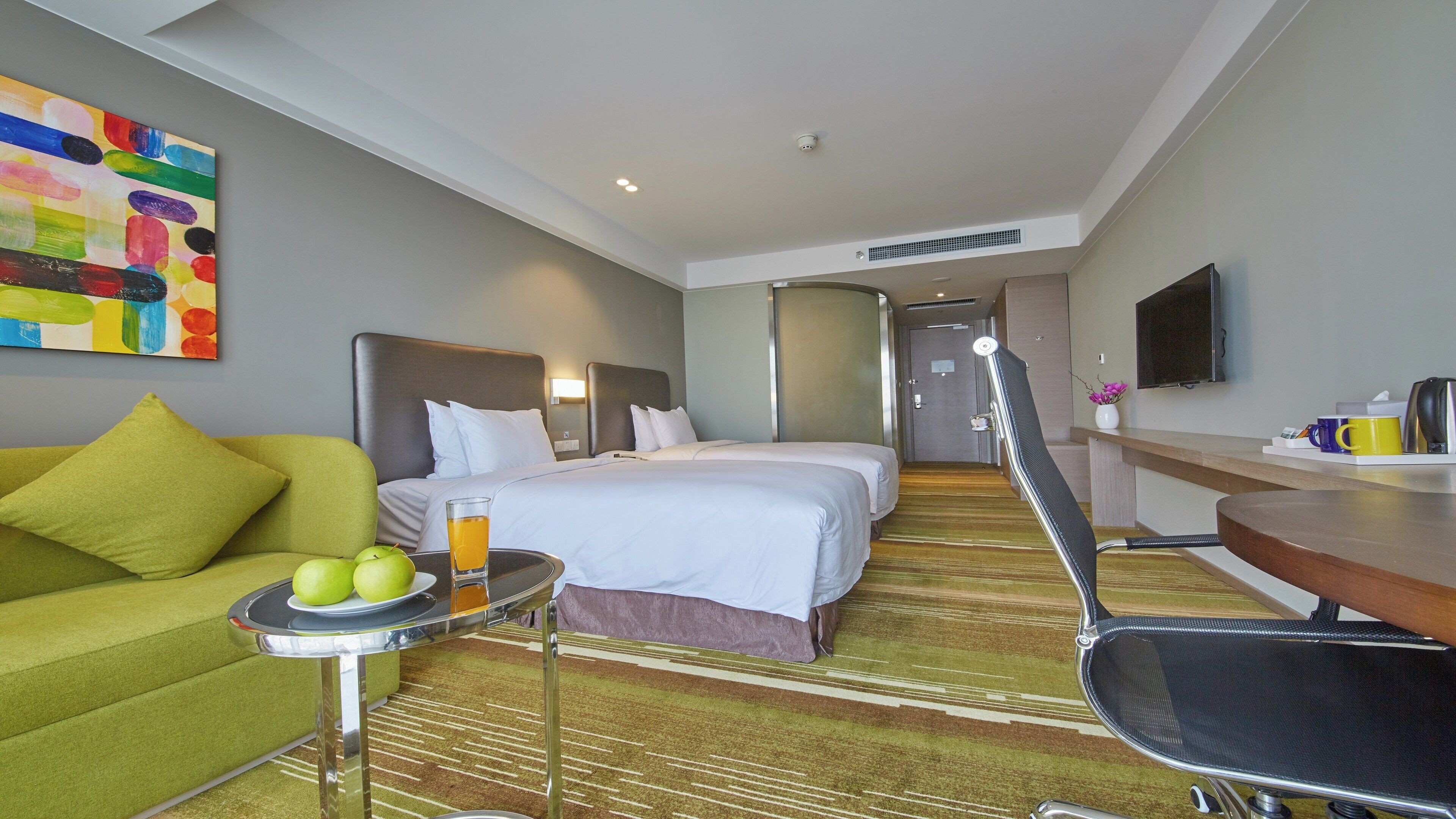 Habitación de hotel con cama, sofá, una mesa con manzanas y una bebida, y un televisor en la pared.
