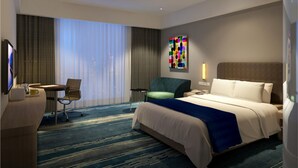 Hotellihuone, jossa on sänky, työpöytä, tuoli, suuri ikkuna verhoilla sekä värikäs taideteos seinällä.