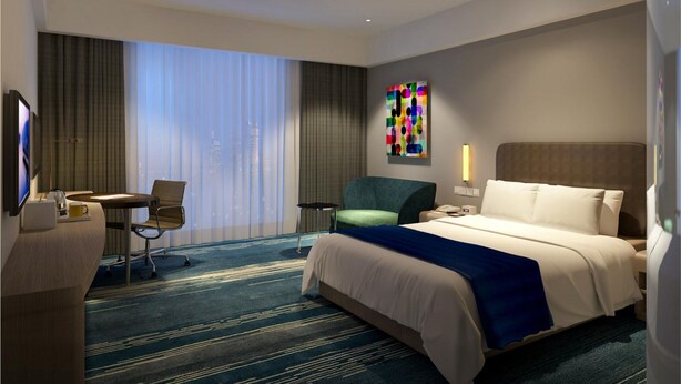 A hotel room with a bed, a desk, a chair, a large window with curtains, and a colorful artwork on the wall.