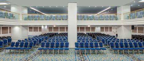 Une grande salle de conférence avec des rangées de chaises bleues, une moquette à motifs et un balcon agrémenté de panneaux de verre décoratifs.