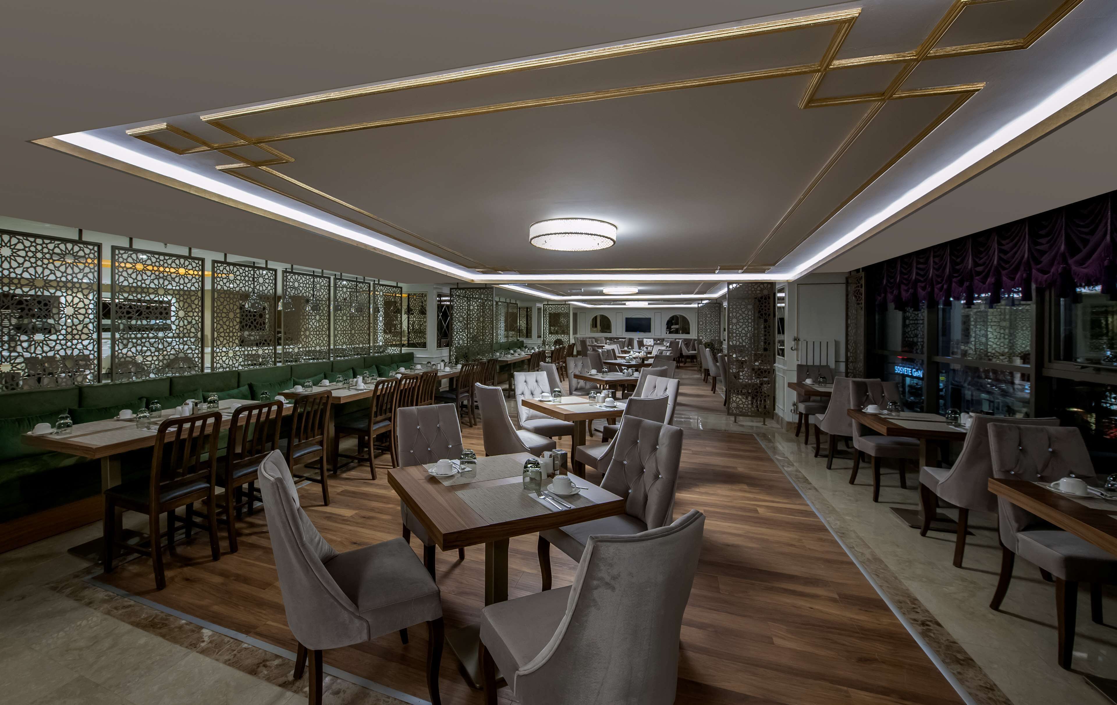 A spacious restaurant with multiple dining areas, a high ceiling with geometric light fixtures, and a view of the city through large windows.