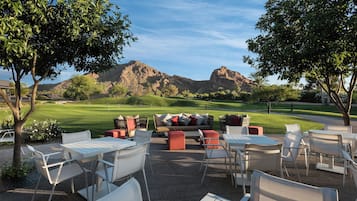 An outdoor seating area with tables and chairs, overlooking a golf course and mountains.