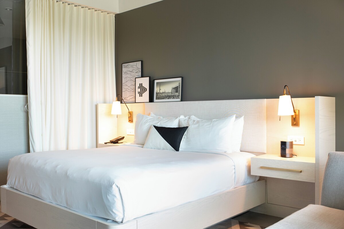 A hotel room with a large bed, two bedside tables, a nightstand, a wall-mounted lamp, and framed pictures.