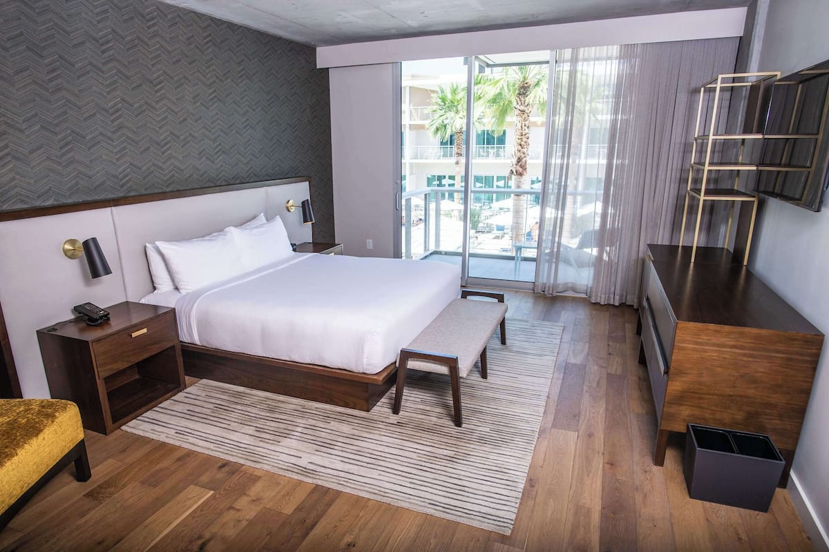 A hotel room with a large bed, a bedside table with a lamp, a bench, a wooden dresser, and a view of the outdoor area.